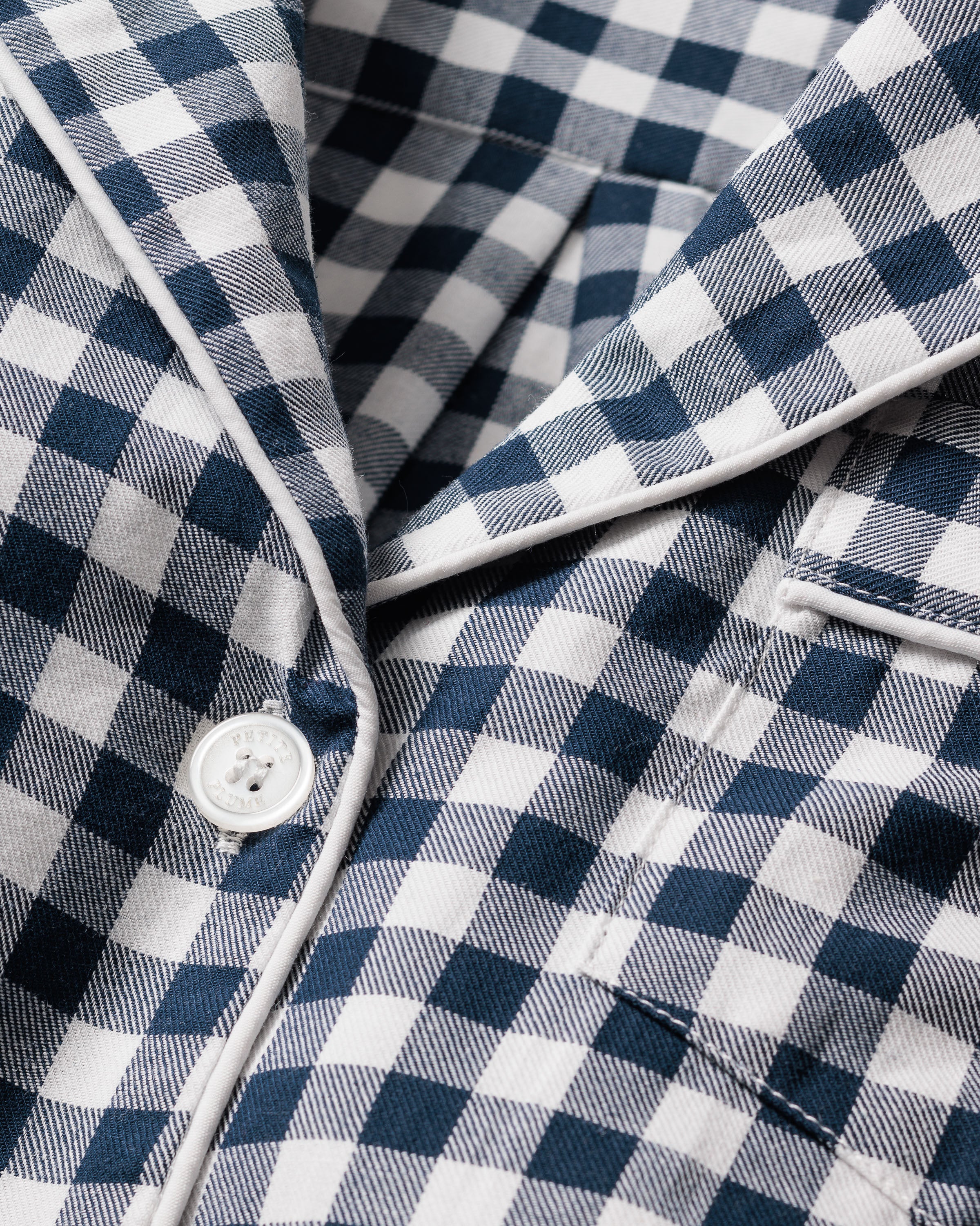 Close-up of the Womens Twill Pajama Set in Navy Gingham by Petite Plume, showcasing a blue and white gingham print with buttons and white piping. The soft cotton twill is neatly stitched, evoking the comfort of pajamas.