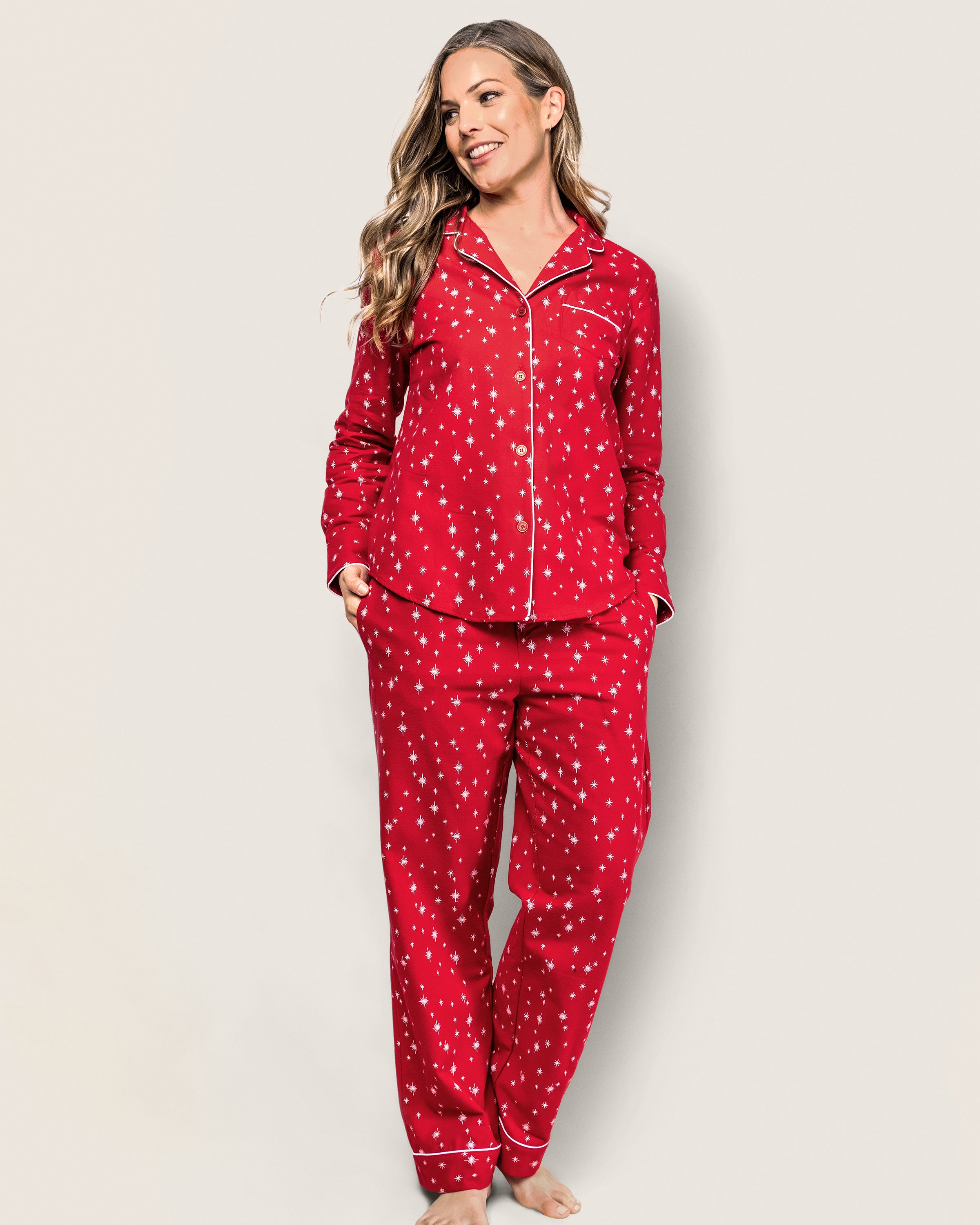 A person in Petite Plumes Womens Flannel Pajama Set in Starry Night, featuring buttons and a collar, stands against a plain backdrop. They smile with long hair, hands cozy in the pockets of the comfy pajama pants adorned with white polka dot-like stars.