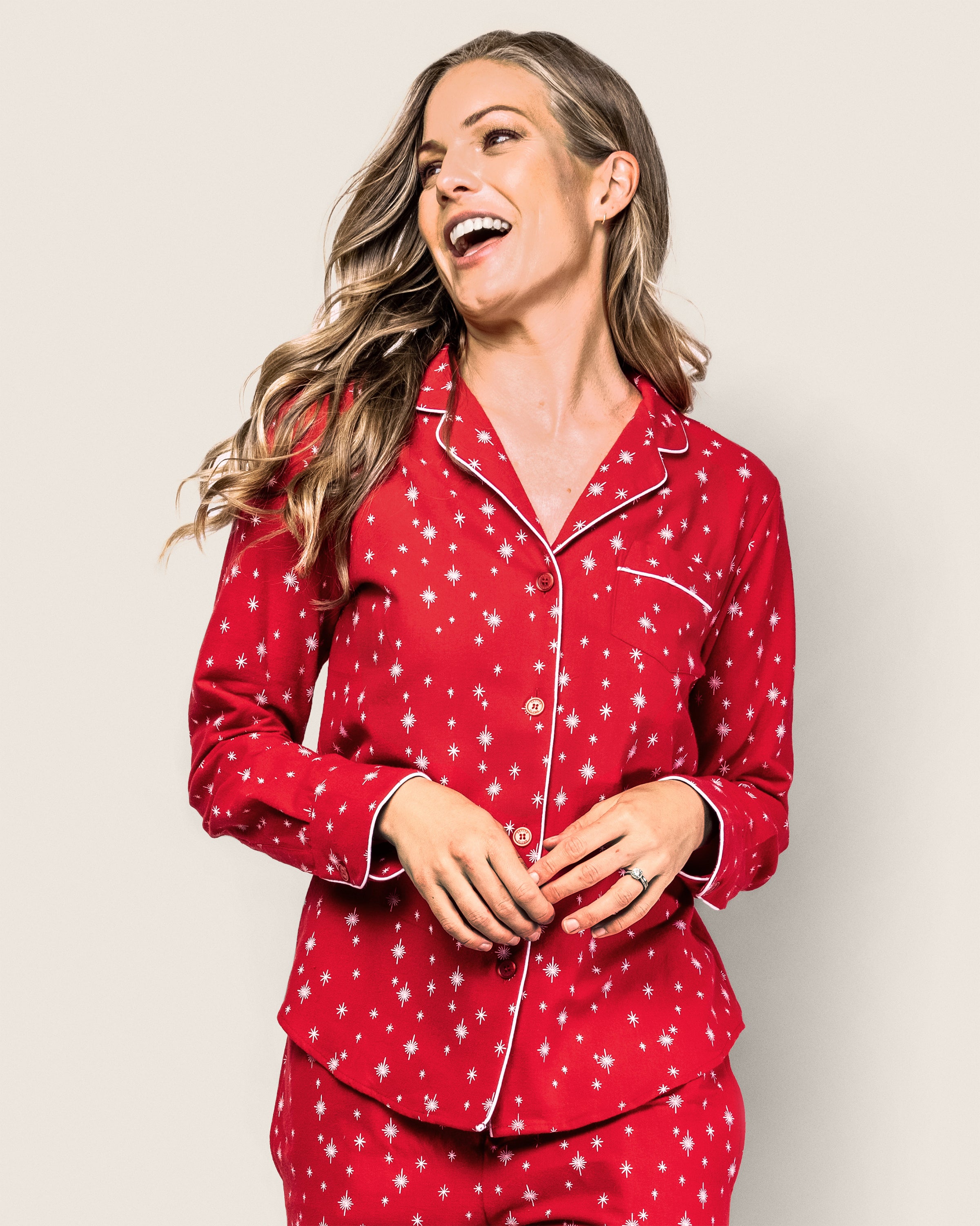 A woman with long wavy hair smiles broadly in her Petite Plume Womens Flannel Pajama Set in Starry Night, adorned with white star patterns and delicate piping. Her hands are clasped gently, adding to the cozy scene against a plain light background.