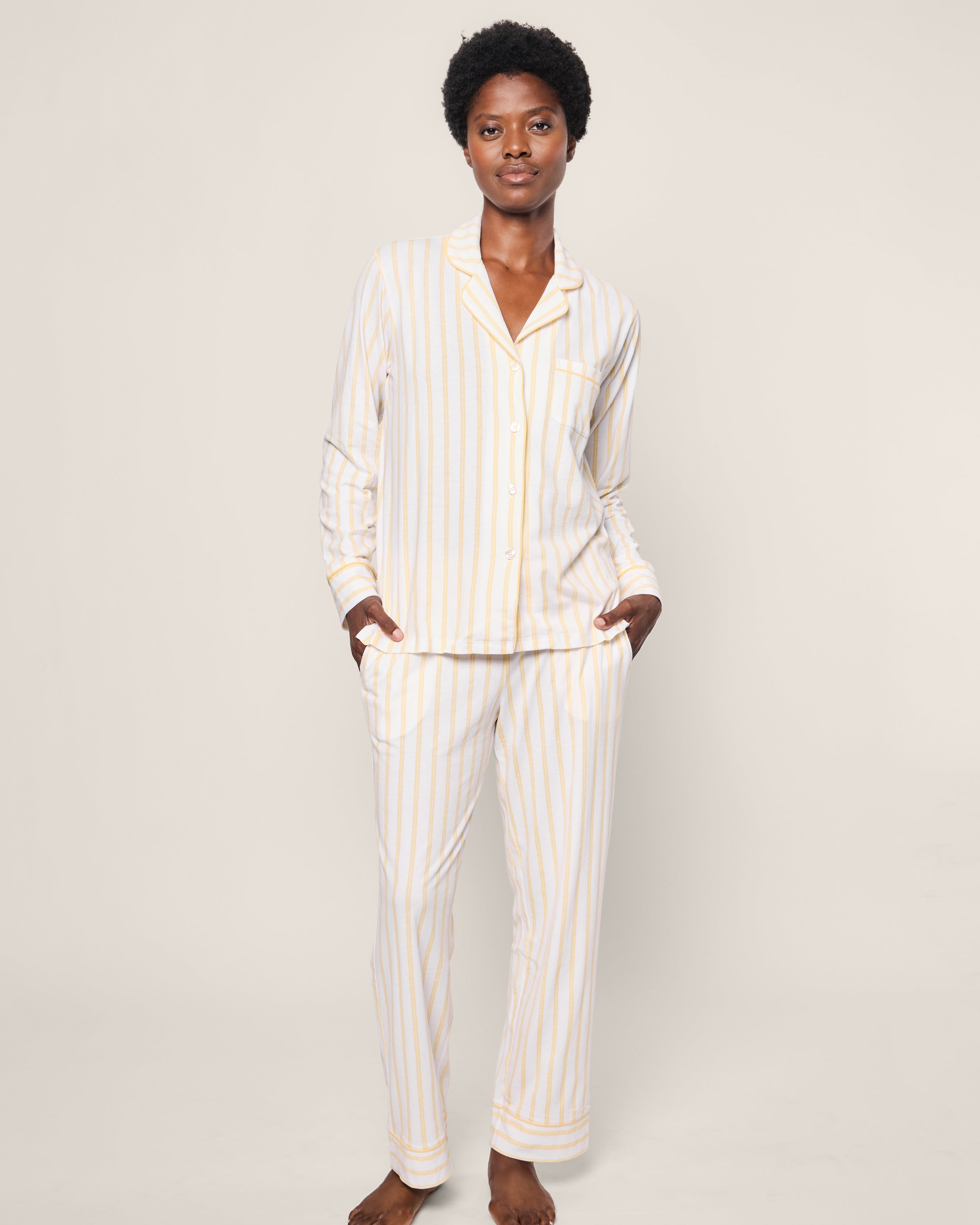 A person wearing Petite Plumes Womens Pima Pajama in Sunny Stripe, made from buttery soft Peruvian Pima cotton, stands against a plain background. The luxury sleepwear set features vertical yellow and white stripes, a button-up shirt with pants, and the person has one hand in their pocket.