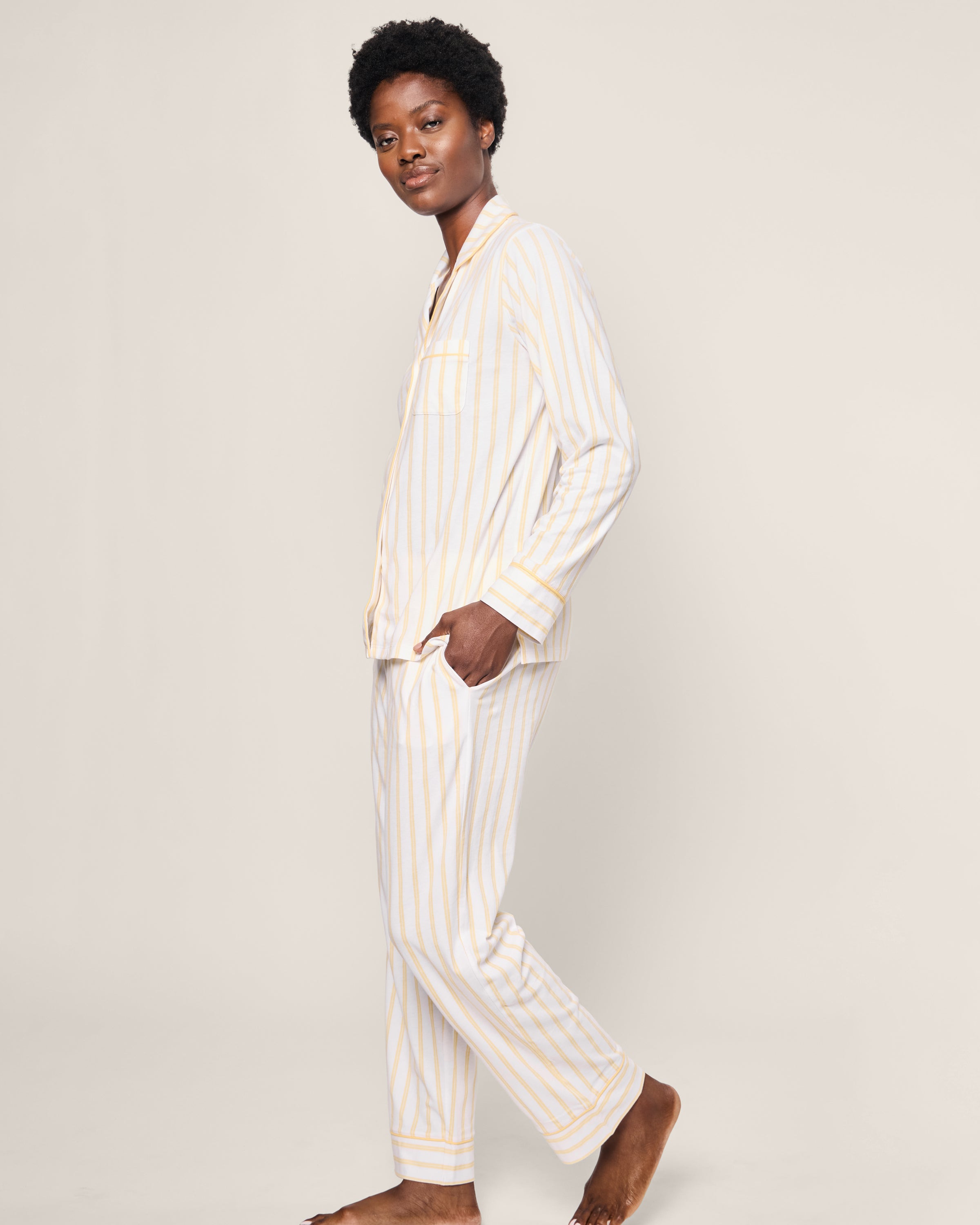 A person wears the Womens Pima Pajama in Sunny Stripe by Petite Plume, featuring buttery soft Peruvian Pima cotton with light-colored stripes, standing sideways with hands in pockets against a plain background.