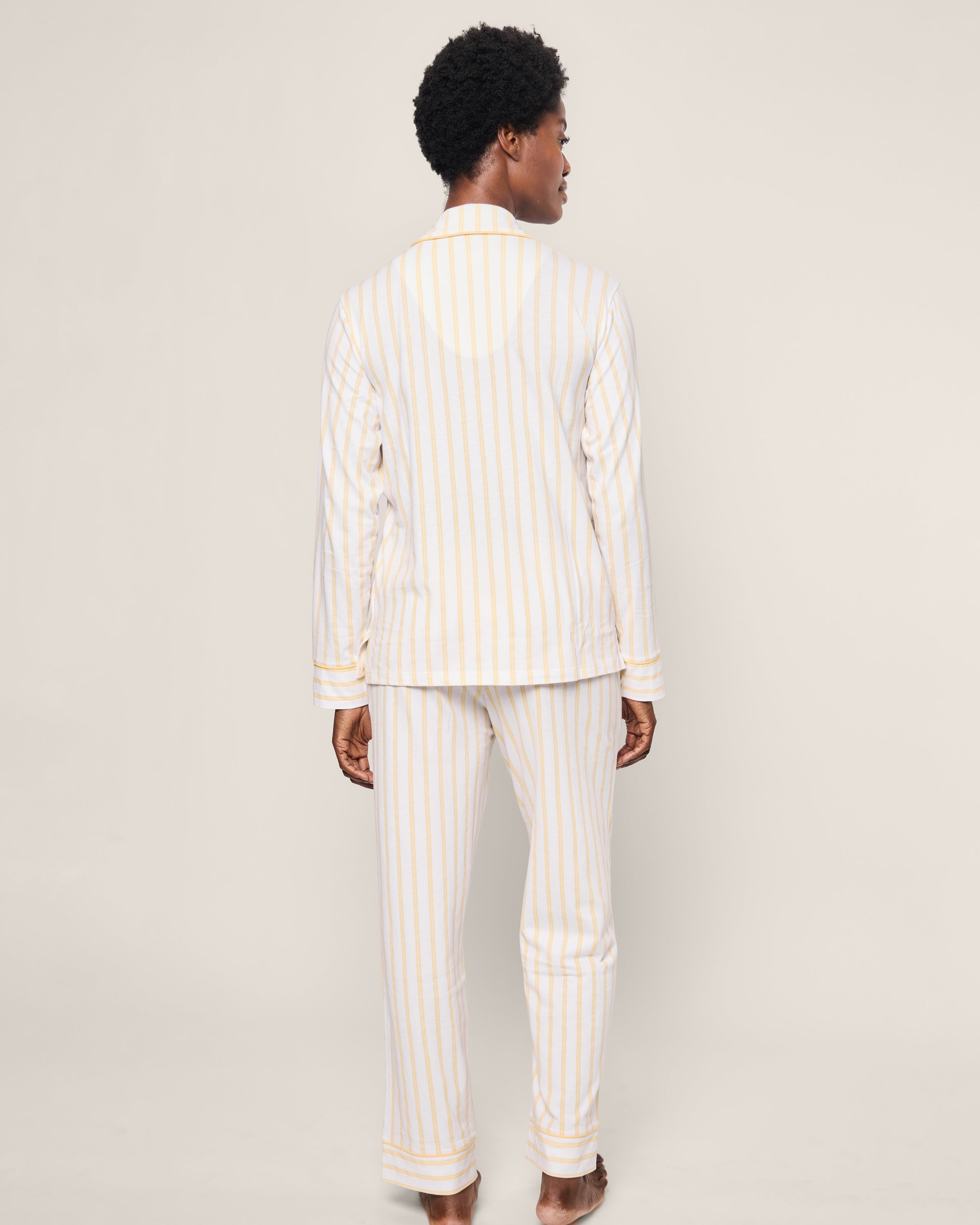 A person is seen from the back in Petite Plumes Womens Pima Pajama in Sunny Stripe, made from buttery soft Peruvian Pima cotton. The luxe sleepwear has light vertical stripes on white, showcasing cozy elegance as the barefoot wearer stands against a plain backdrop.