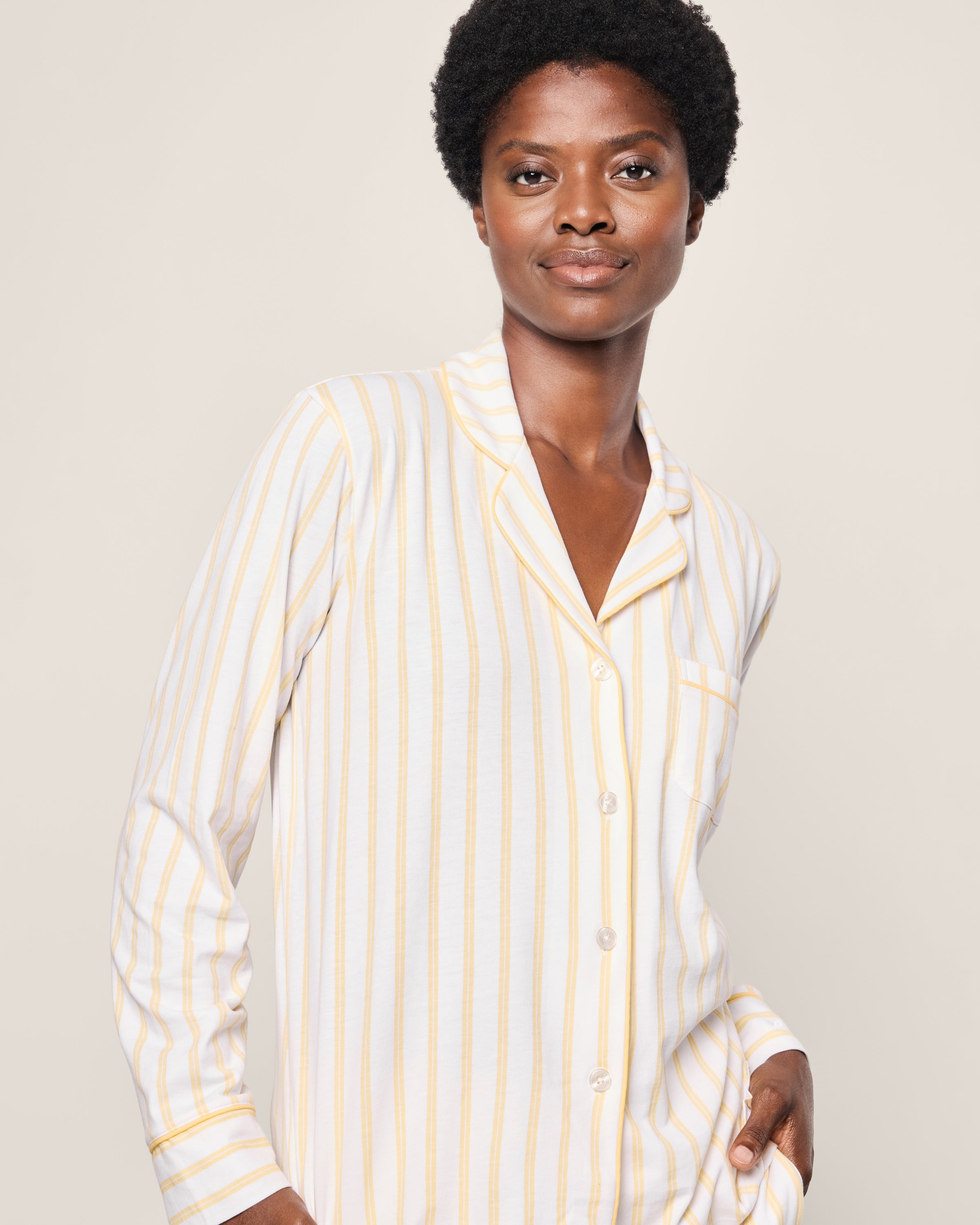 A person in a Petite Plume Womens Pima Pajama in Sunny Stripe, featuring light-colored yellow stripes on white Peruvian Pima cotton. They have short curly hair, gaze into the camera with a slight smile against a plain background.