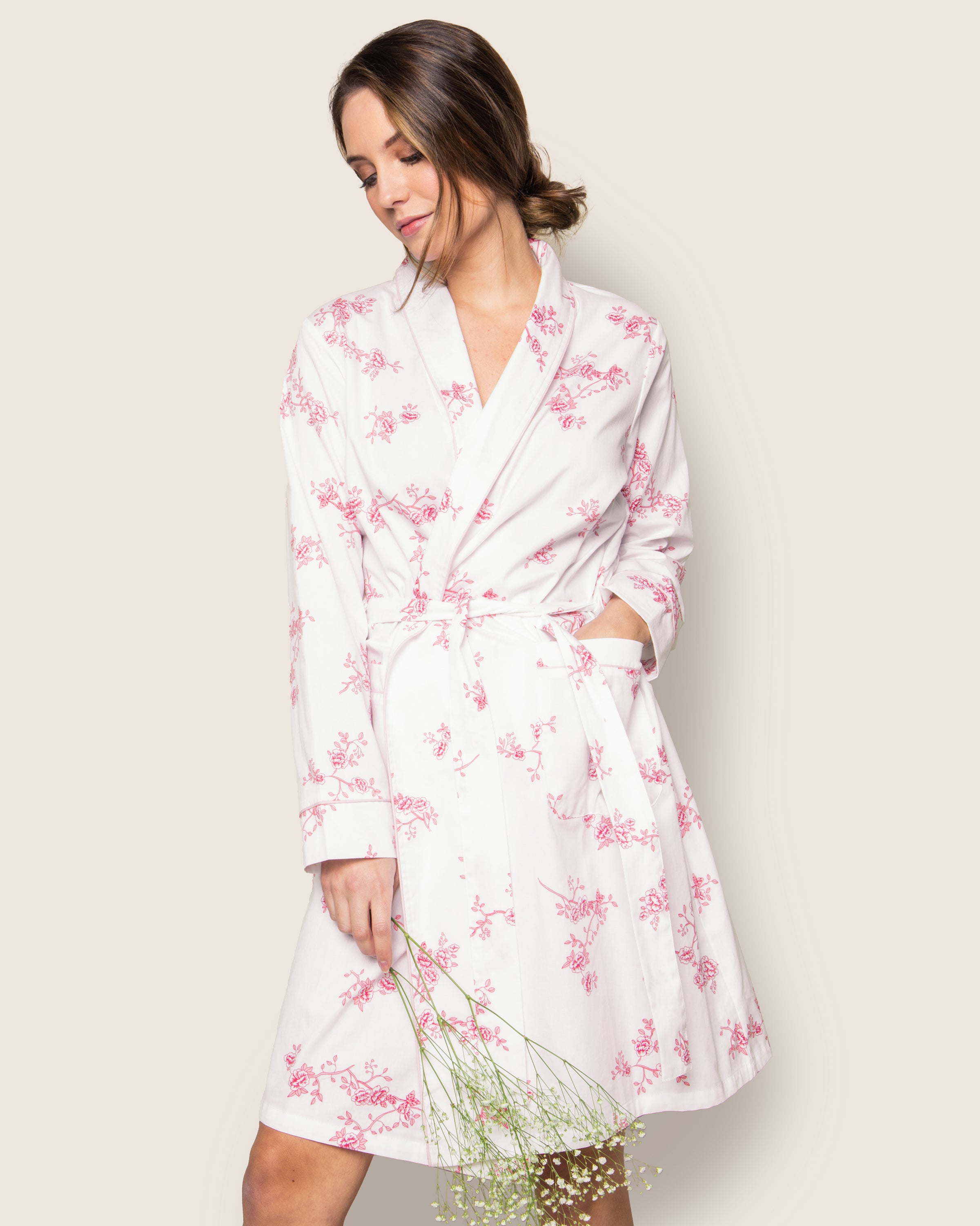 A woman stands relaxed against a neutral background, wearing Petite Plumes Womens Twill Robe in English Rose Floral. This luxurious robe, featuring pink floral patterns on quality cotton sleepwear with long sleeves and a matching belt, complements the small white flowers she holds.