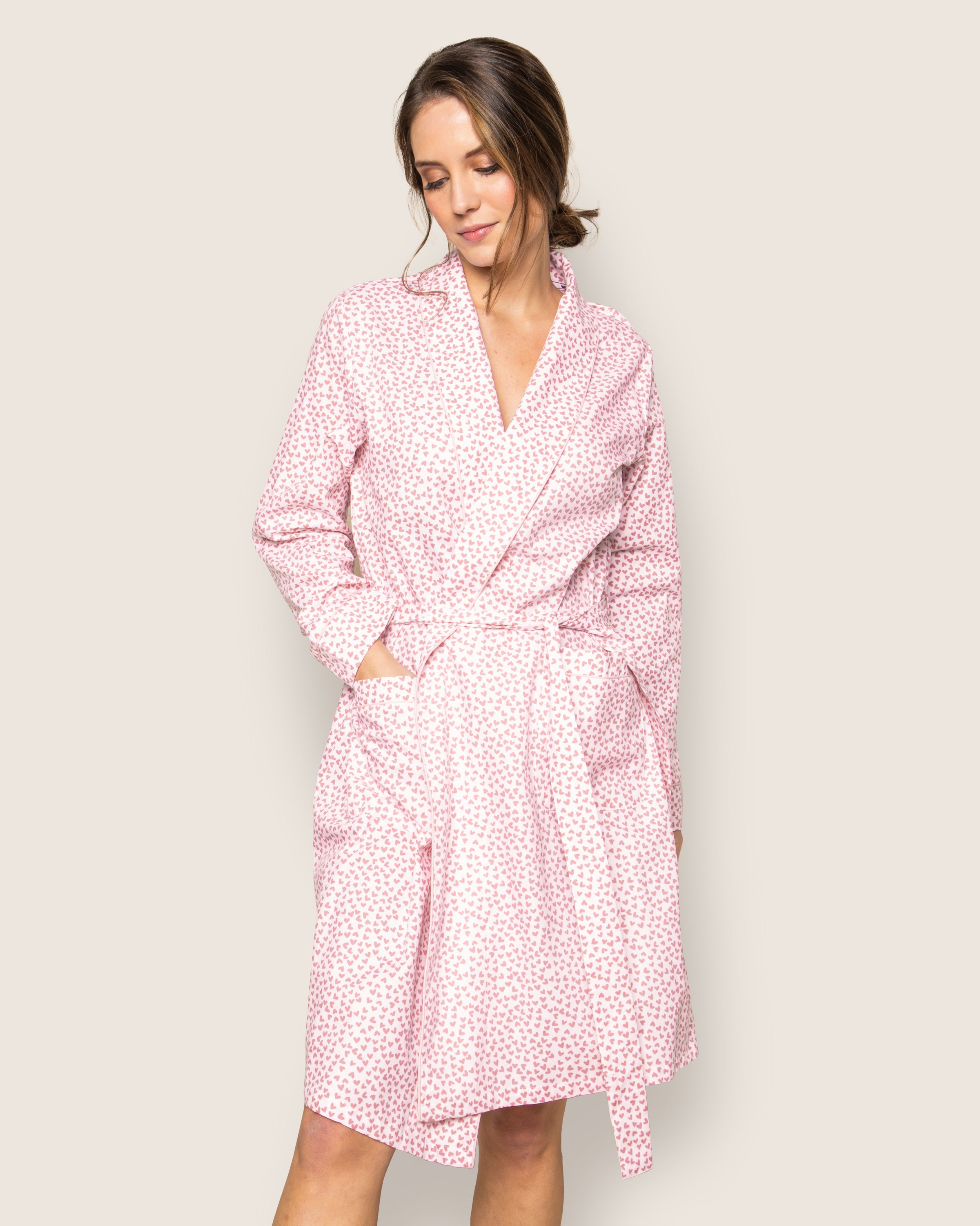 A woman in a Womens Twill Robe in Sweethearts by Petite Plume poses against a neutral background. Her left hand rests in the robes pocket, her hair styled in a low bun, and she smiles gently, embodying luxurious sleepwear.