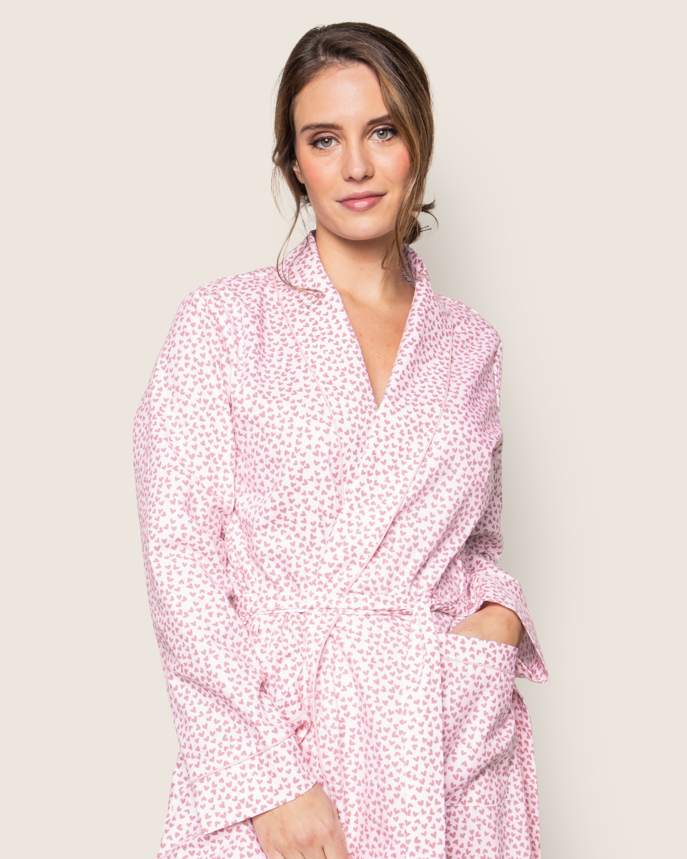 A person with brown hair wears the Womens Twill Robe in Sweethearts by Petite Plume. Crafted from top-quality cotton and adorned with heart patterns, they pose against a neutral background, one hand in a pocket, exuding effortless charm in their luxurious sleepwear.