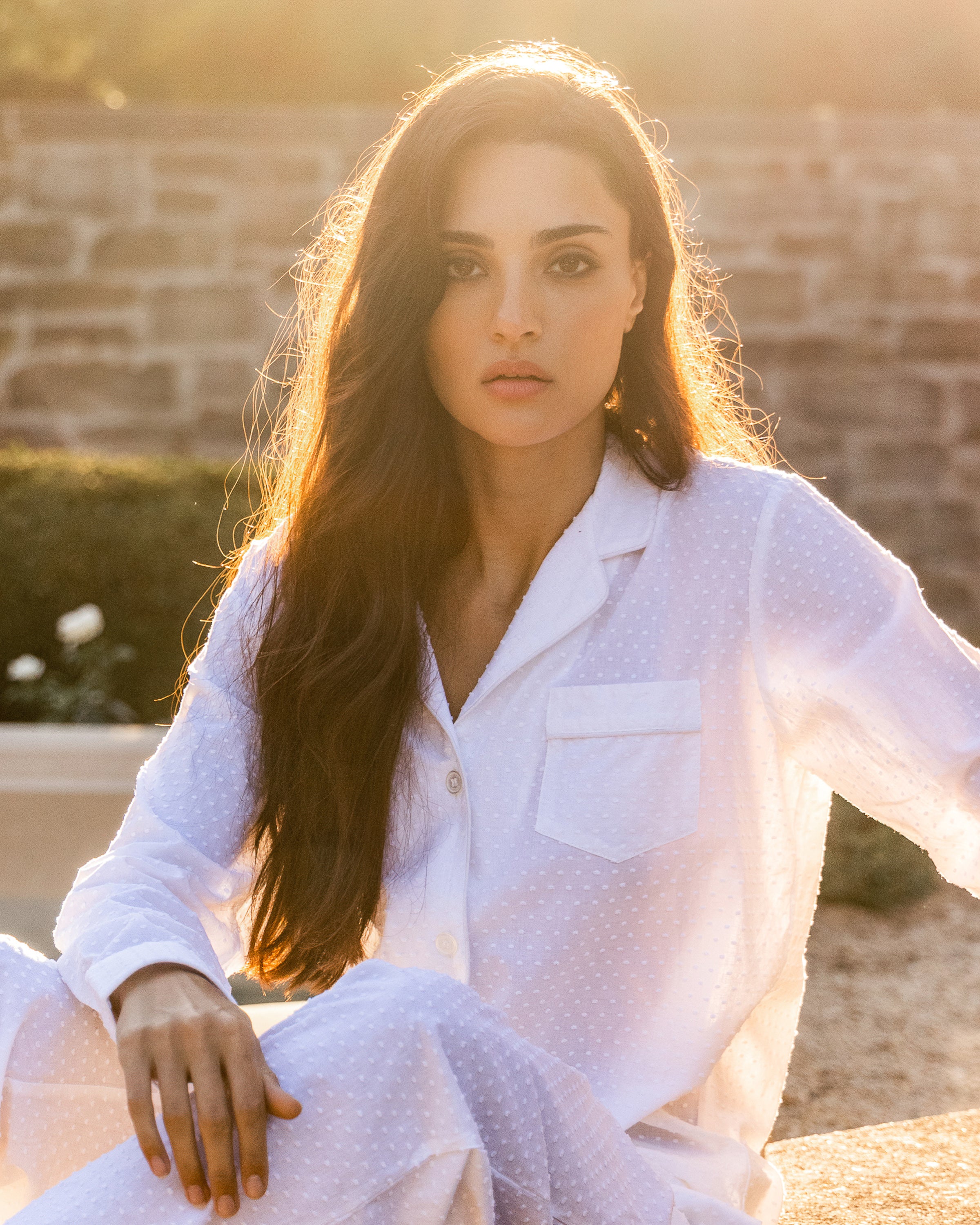 A person with long, dark hair is outdoors in the luxurious Womens Swiss Dots Pajama Set in White by Petite Plume. Sunlight creates a soft glow behind them as they sit seriously on a low wall or platform.