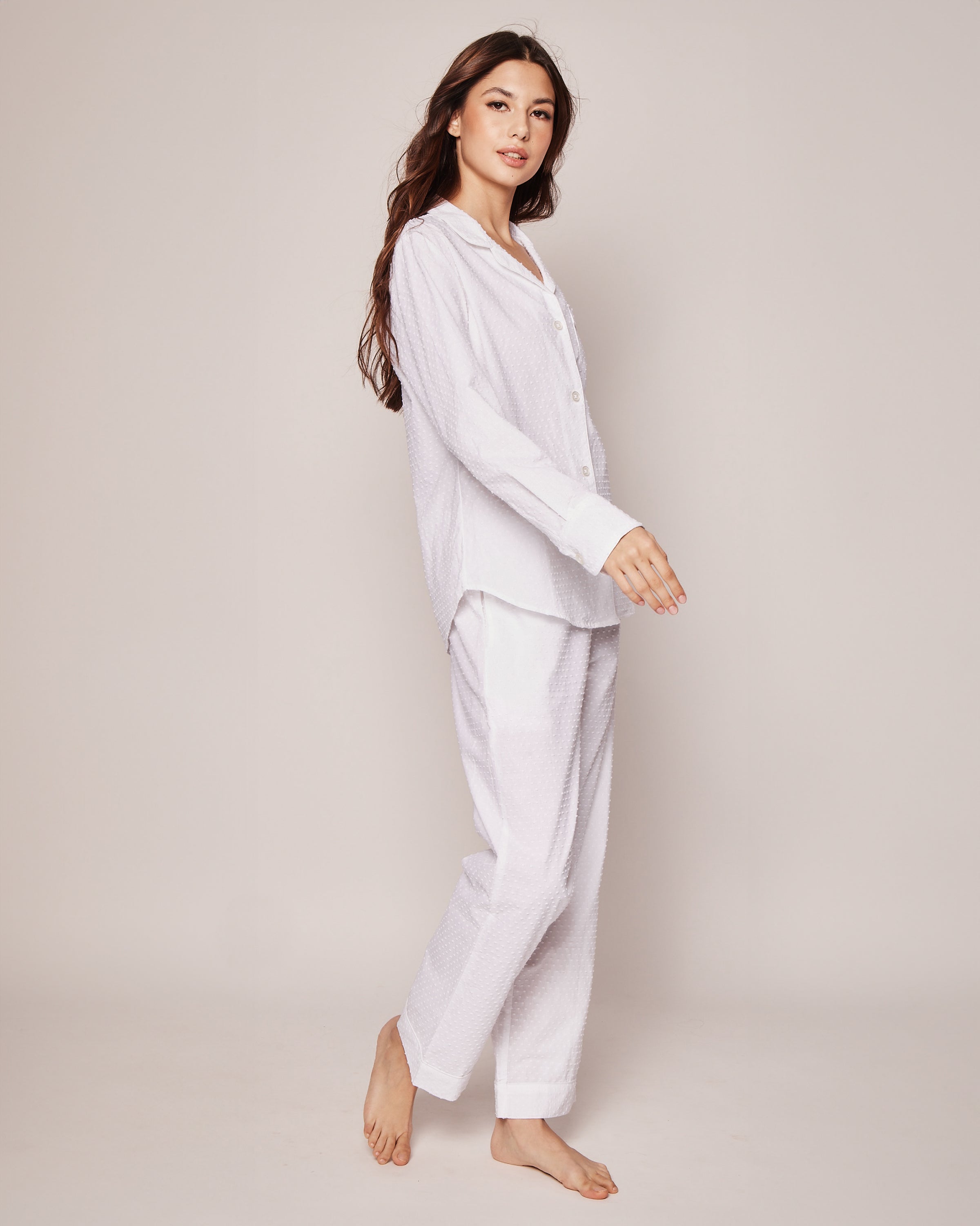 The woman, barefoot against a light background with her long hair cascading, exudes effortless summer charm in the Womens Swiss Dots Pajama Set by Petite Plume. The luxurious set includes a long-sleeved top and pants in classic Cotton Swiss Dot.