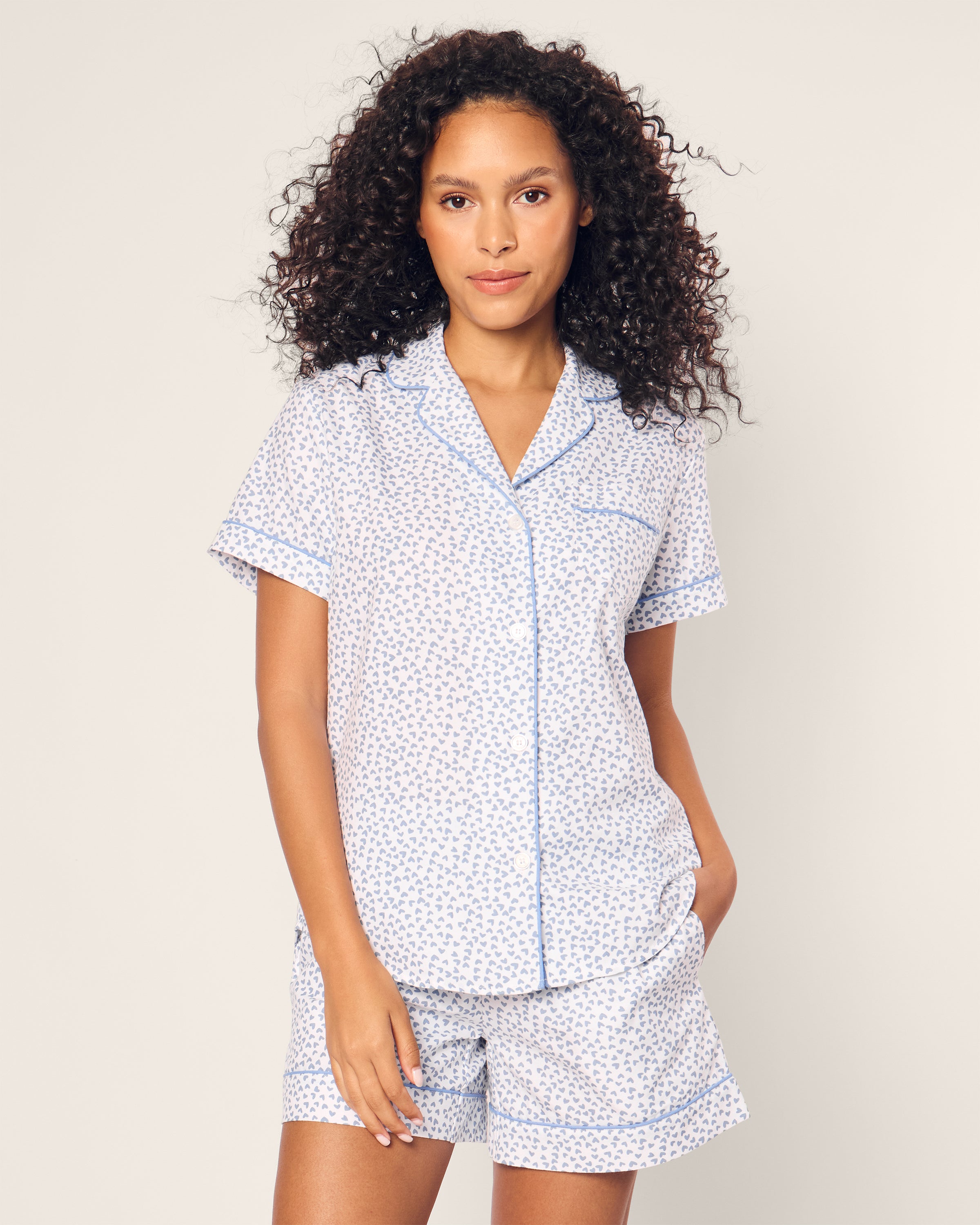 A curly-haired person wears the luxury Petite Plume Womens Twill Short Sleeve Short Set in Bluehearts, featuring a small floral pattern on a light background. The collared top and comfy shorts complement each other perfectly.