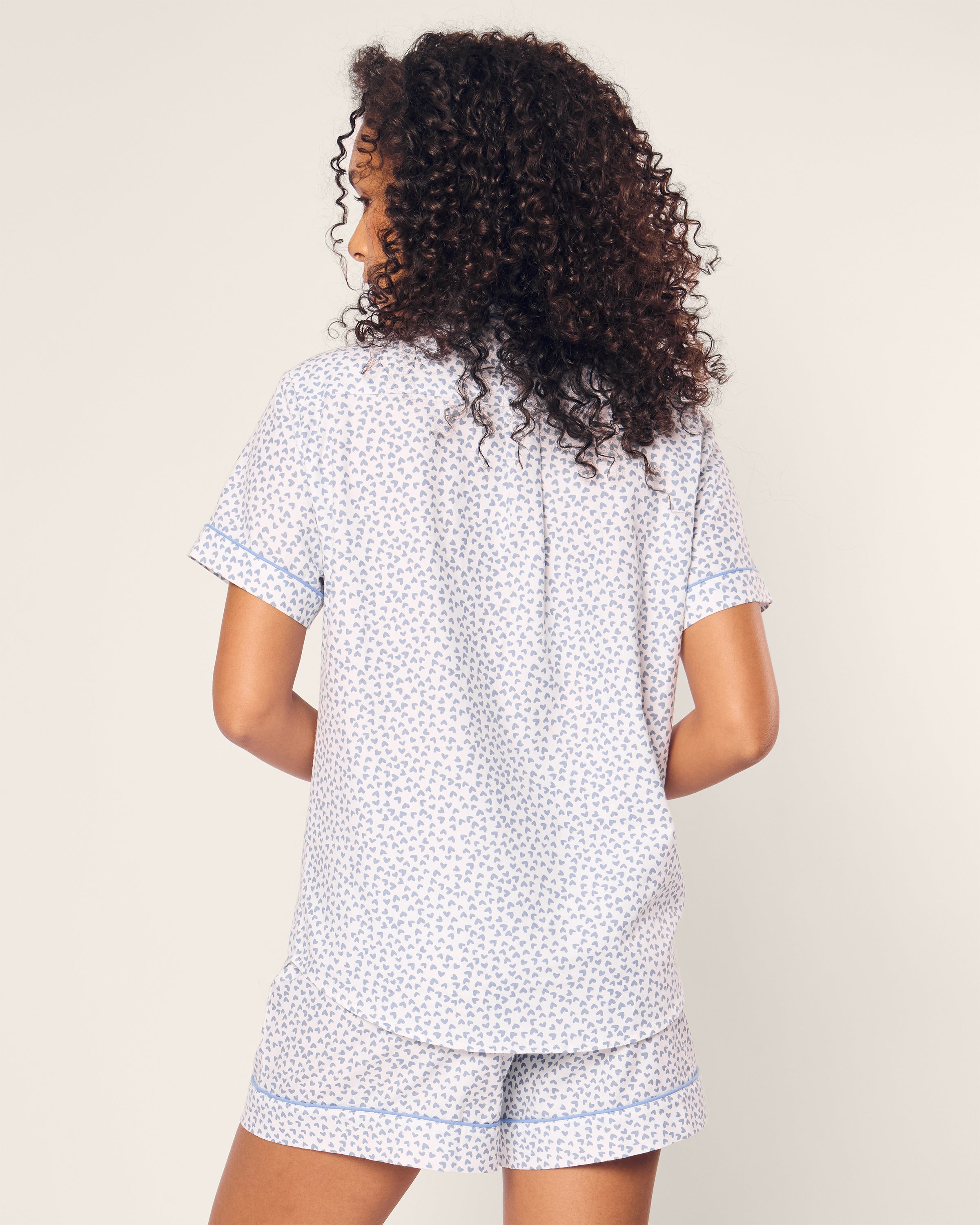 A curly-haired person wears Petite Plumes Womens Twill Short Sleeve Short Set in Bluehearts, featuring a luxury cotton pajama with shorts and a short-sleeved shirt. They face away from the camera against a plain background.