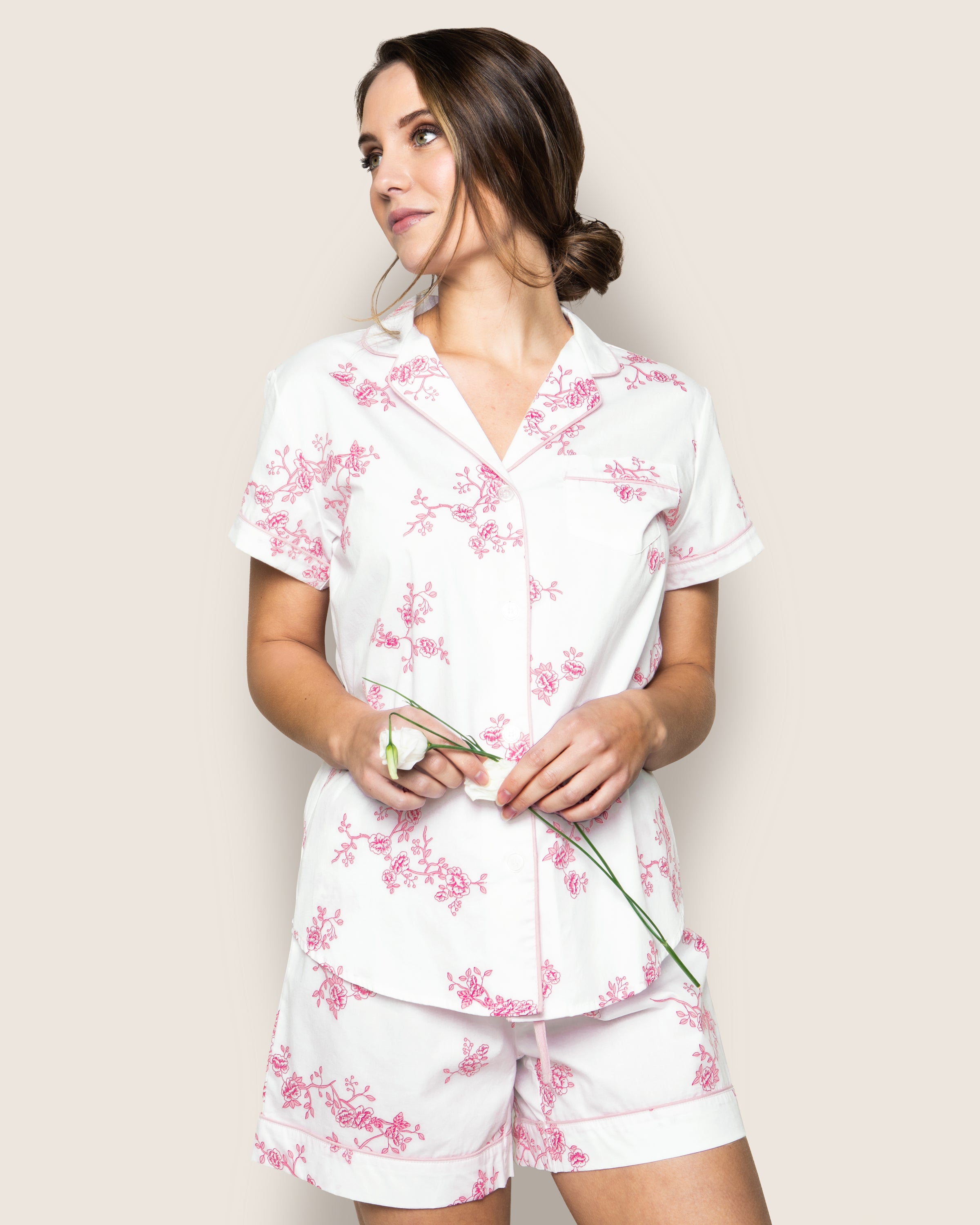 Wearing Petite Plumes Womens Twill Pajama Short Set in English Rose Floral, a person with a low bun stands against a plain background, holding a single white flower and gazing to the side.