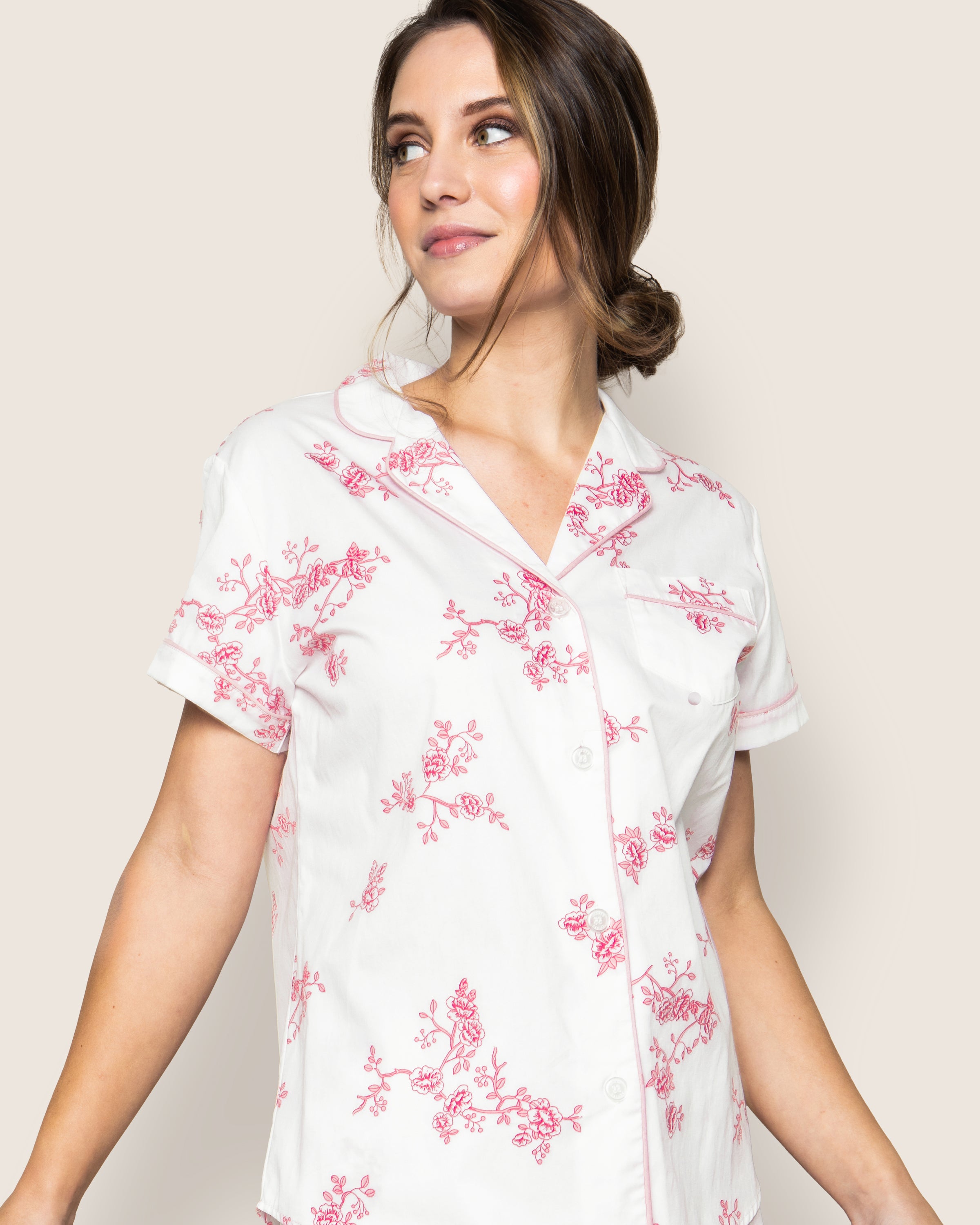 A woman wearing the Womens Twill Pajama Short Set in English Rose Floral by Petite Plume stands against a light background. Her hair is in a low bun, and she gazes to the side with a relaxed expression, embodying the elegance of luxury sleepwear.