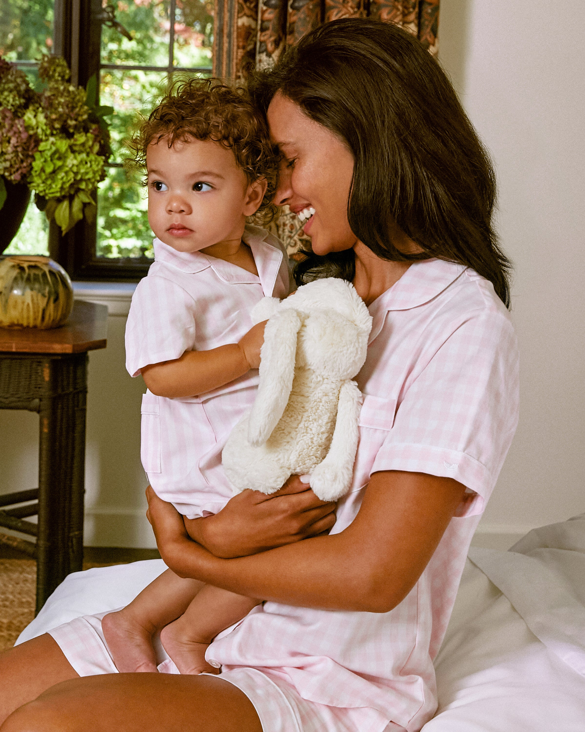 In a cozy room filled with sunlight and plants, a woman in Petite Plumes Womens Twill Pajama Short Set in Pink Gingham cradles a toddler wearing matching pajamas. The toddler cuddles a stuffed bunny, creating a serene and comforting scene.