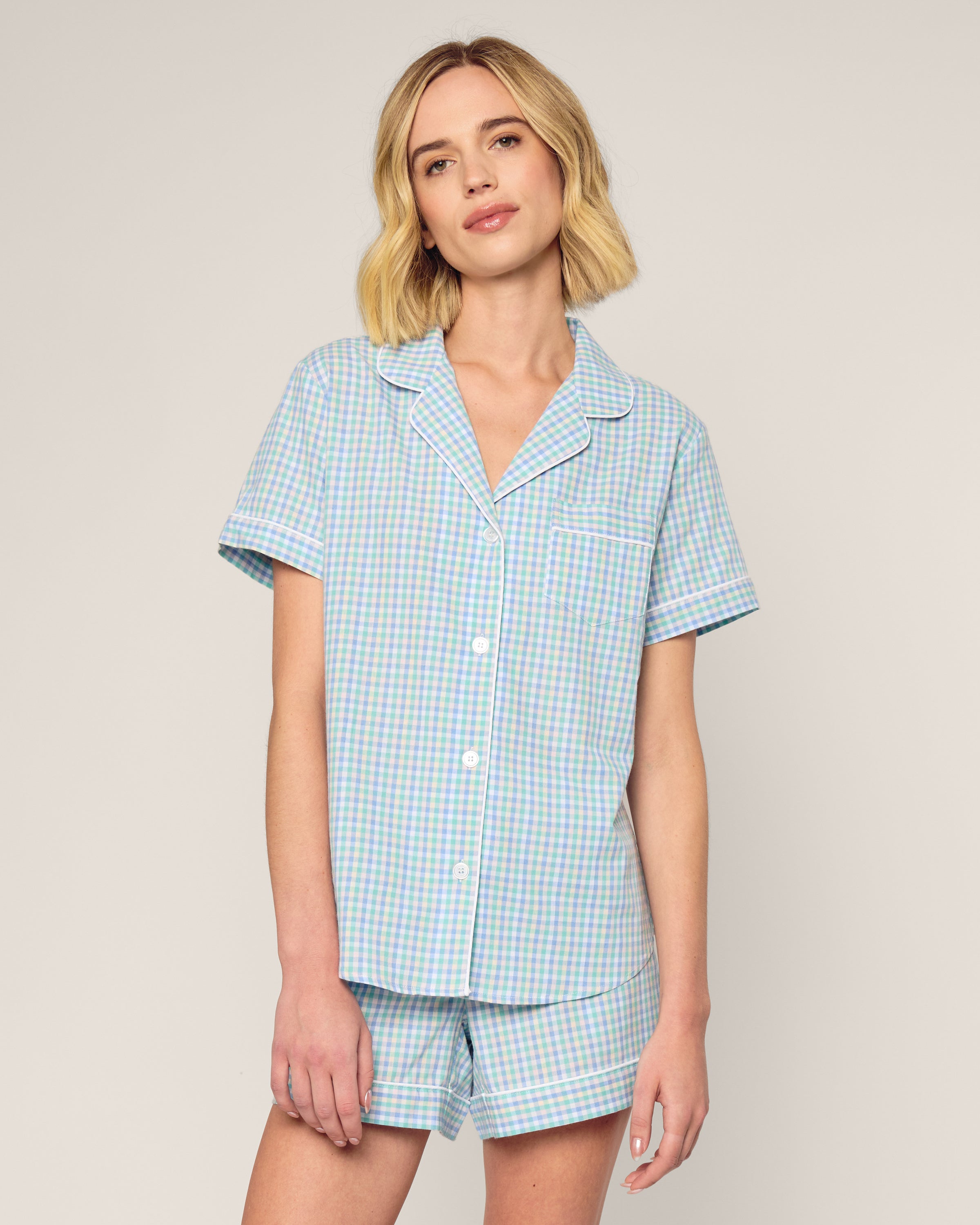 A person with short blonde hair wears the Womens Twill Short Sleeve Short Set in Spring Gingham by Petite Plume, featuring a luxury blue and green gingham button-up shirt and shorts made from soft cotton. They stand against a plain light background.