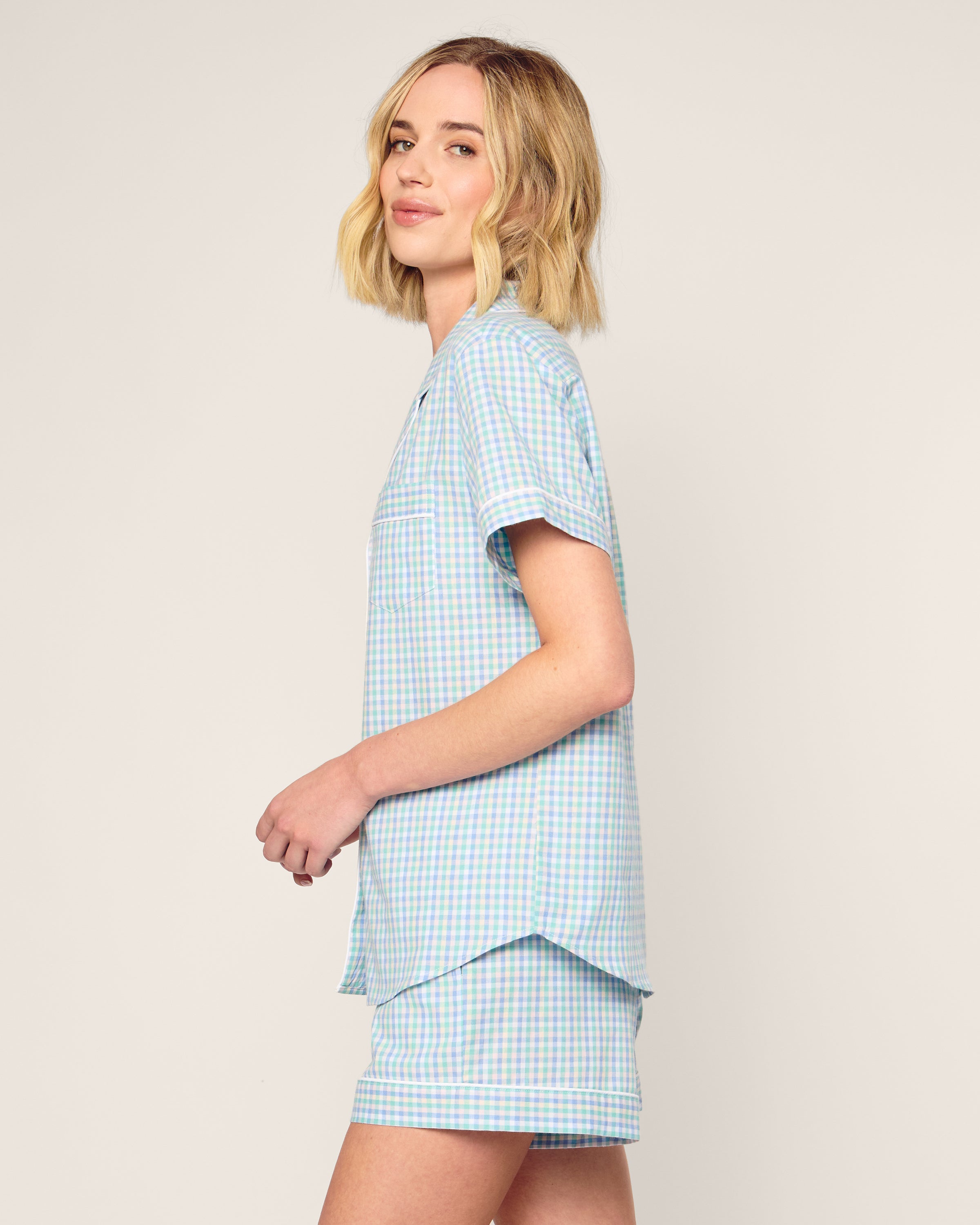 A person with shoulder-length blonde hair is wearing the Womens Twill Short Sleeve Short Set in Spring Gingham by Petite Plume, crafted from soft cotton. They stand sideways, smiling against a plain background, exuding relaxed luxury sleepwear charm.