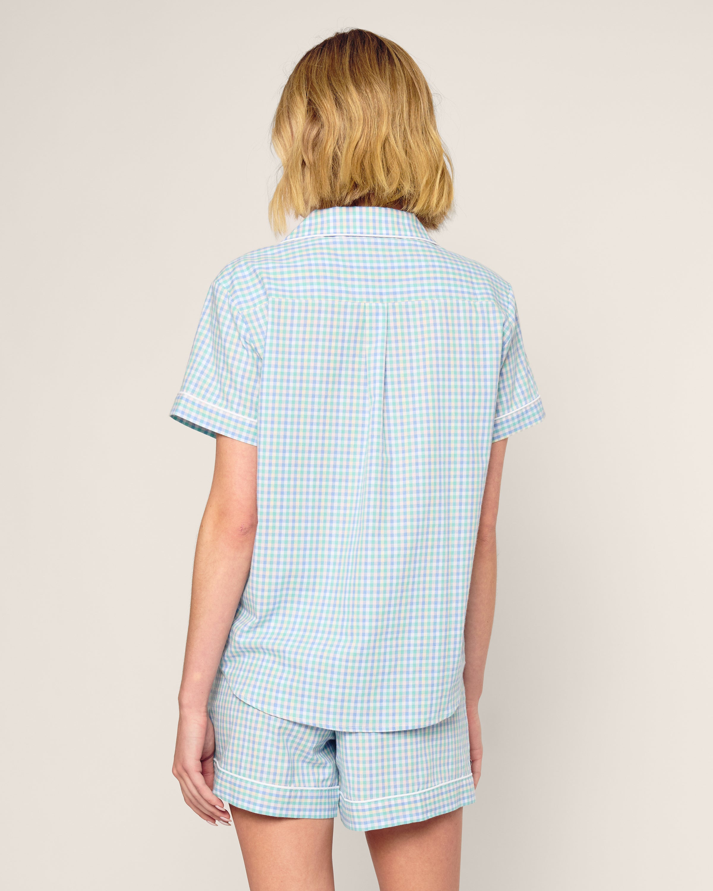 A person with shoulder-length blonde hair wears the Womens Twill Short Sleeve Short Set in Spring Gingham by Petite Plume, against a plain light beige background, exuding casual charm.