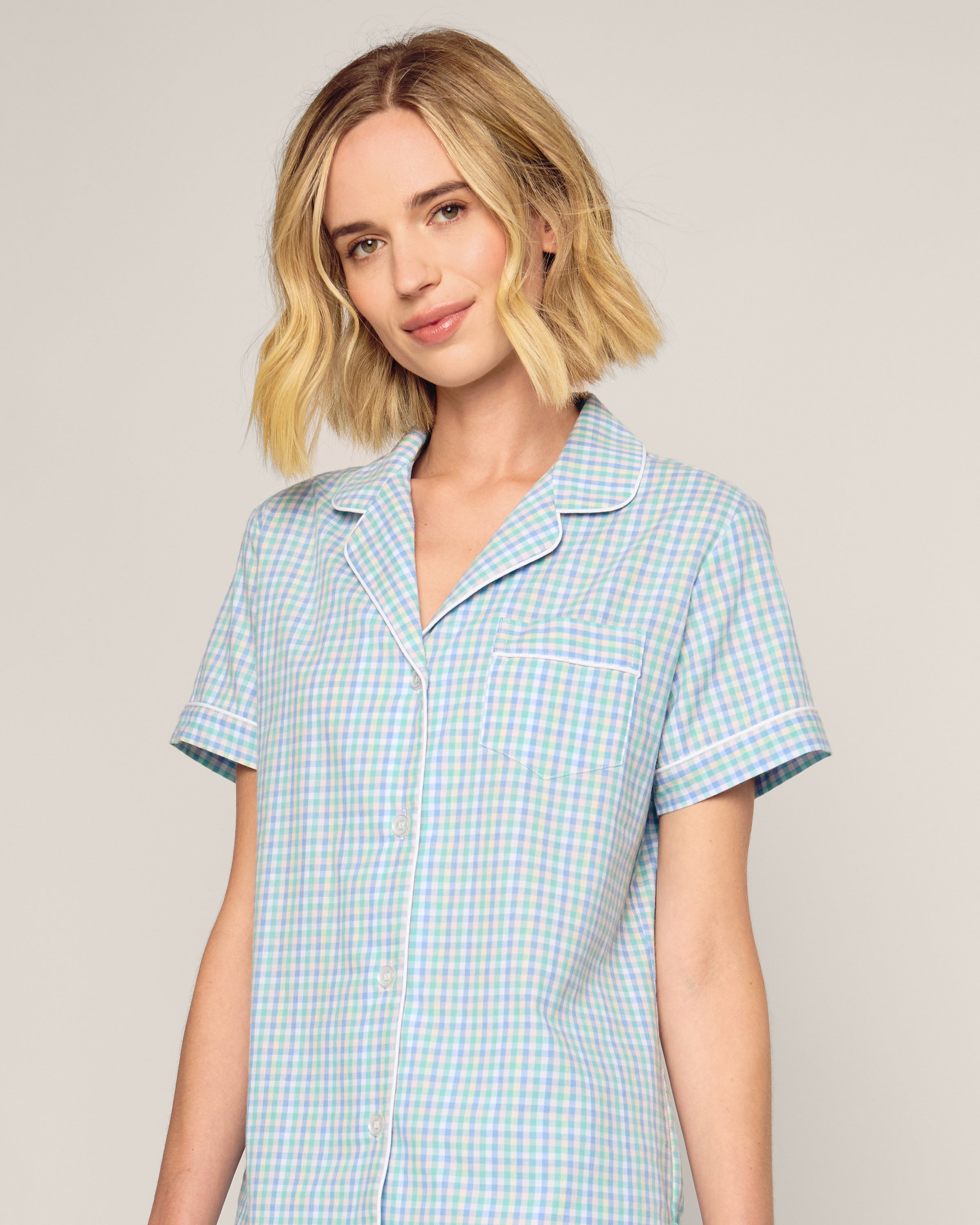 A person with short blonde hair wears Petite Plumes Womens Twill Short Sleeve Short Set in Spring Gingham—a luxury sleepwear set in cotton. Against a plain light background, they smile softly, exuding comfort.