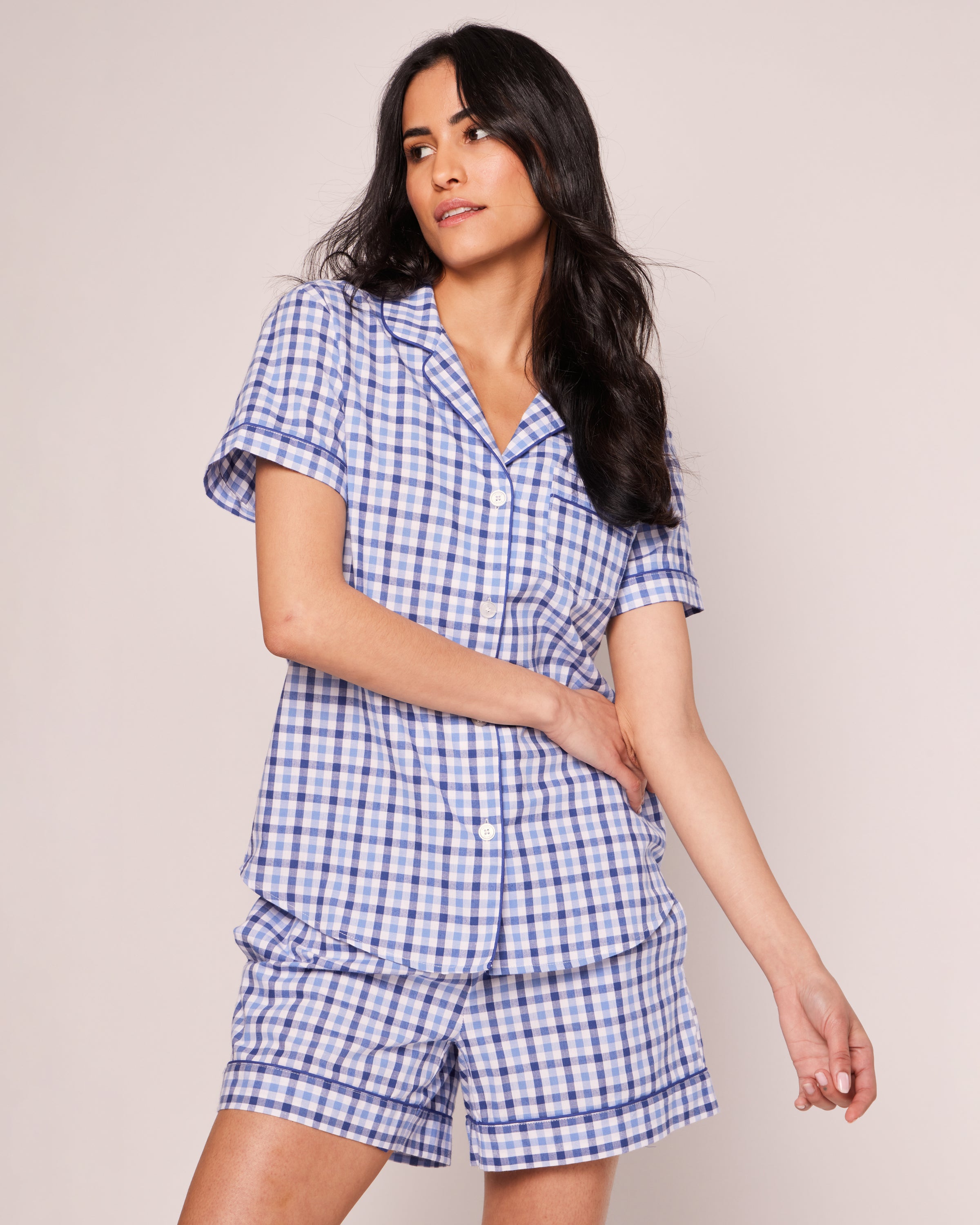 A person with long dark hair wearing Petite Plumes Womens Twill Pajama Short Sleeve Short Set in Royal Blue Gingham poses stylishly, one hand on their waist and the other extended, against a plain off-white background, capturing the luxury of sleepwear.