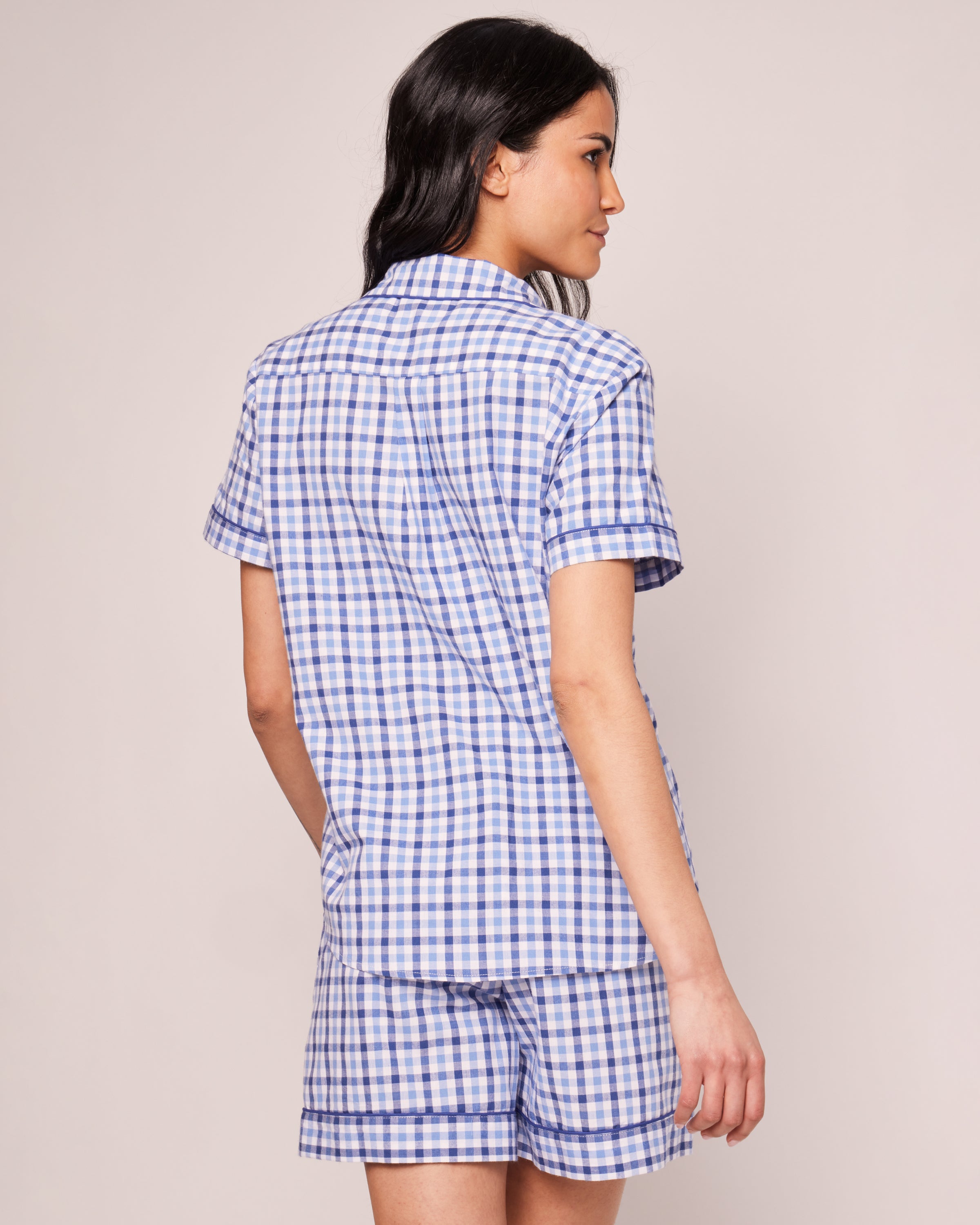 A woman with long dark hair stands against a plain background, wearing the Petite Plume Womens Twill Pajama Short Sleeve Short Set in Royal Blue Gingham. She gazes to the side, effortlessly embodying the charm of this luxury sleepwear ensemble.