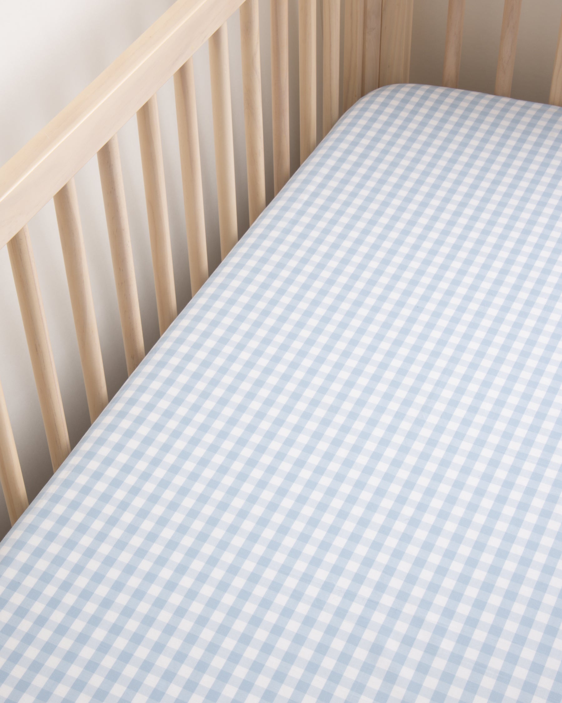 Close-up of a crib with a light wood frame featuring Petite Plumes Luxe Premium Light Blue Gingham Crib Sheet. The ultra-soft cotton percale sheet in blue-and-white checks rests on the mattress, while breathable vertical wooden slats of the crib create a serene nursery ambiance.