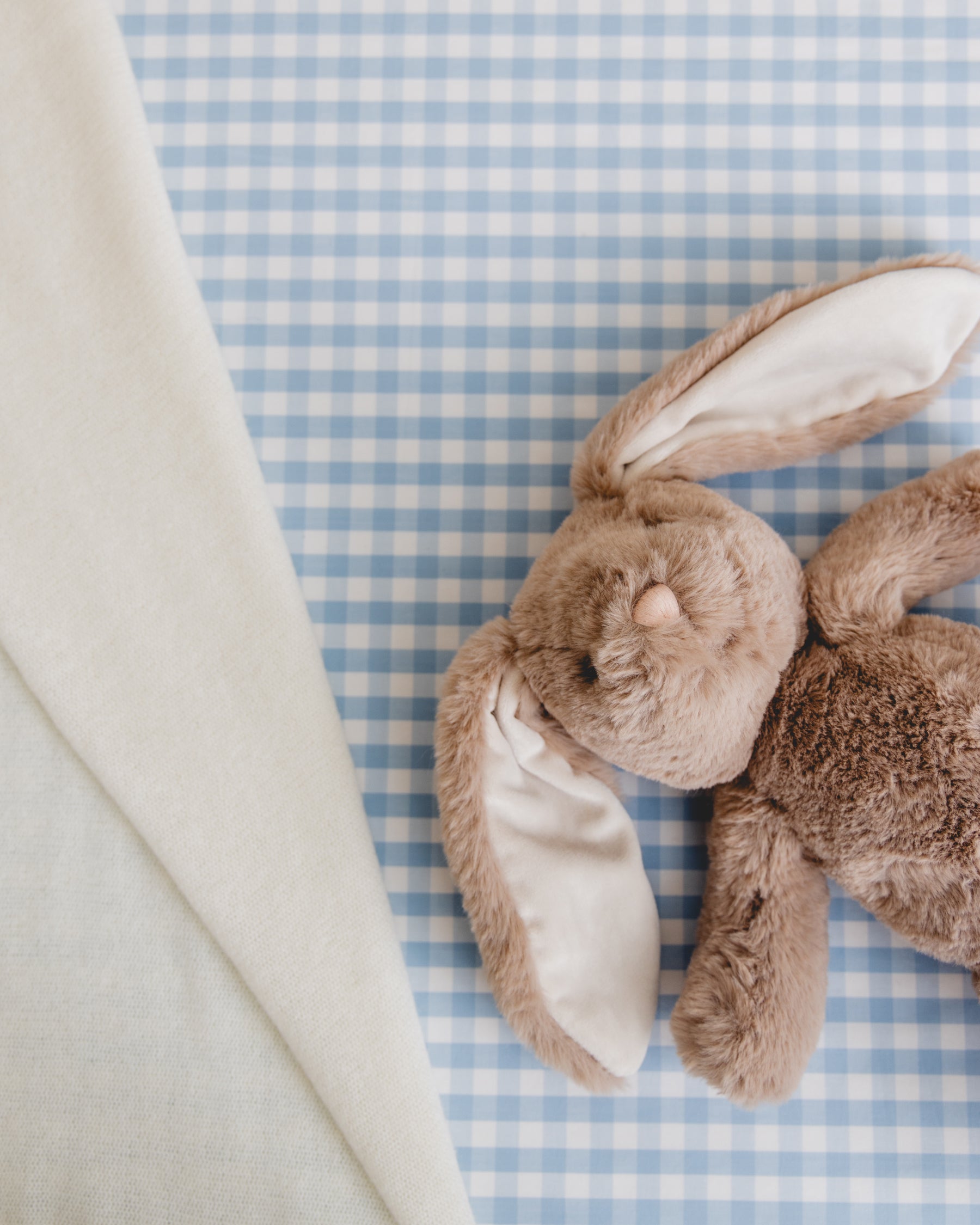 A soft brown plush bunny with floppy ears rests on the Luxe Premium Light Blue Gingham Crib Sheet by Petite Plume, made from breathable cotton percale. A corner of a cream-colored cloth is visible on the left side of the image.