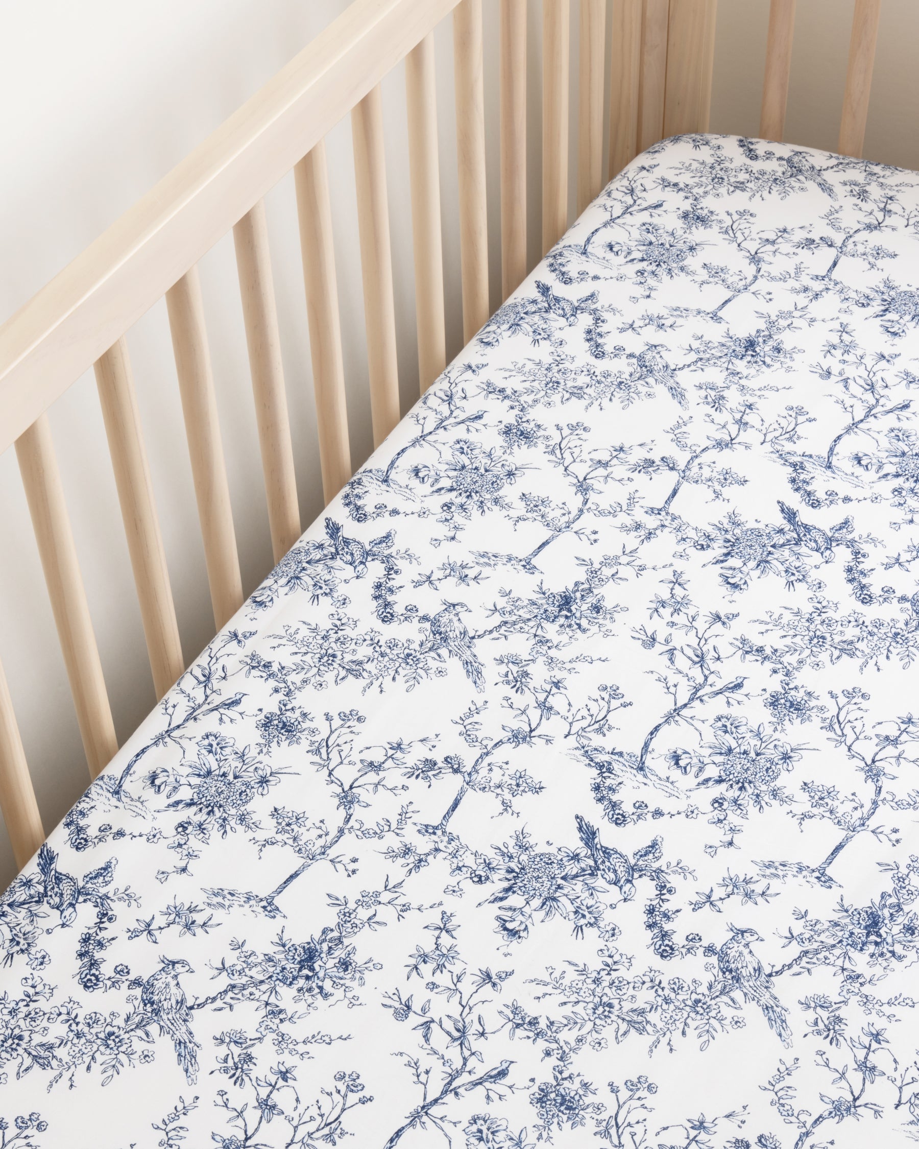 A wooden crib with Luxe Premium Cotton Crib Sheets by Petite Plume, featuring a blue floral pattern. The soft, breathable mattress is encased against horizontal slats and a light wall.