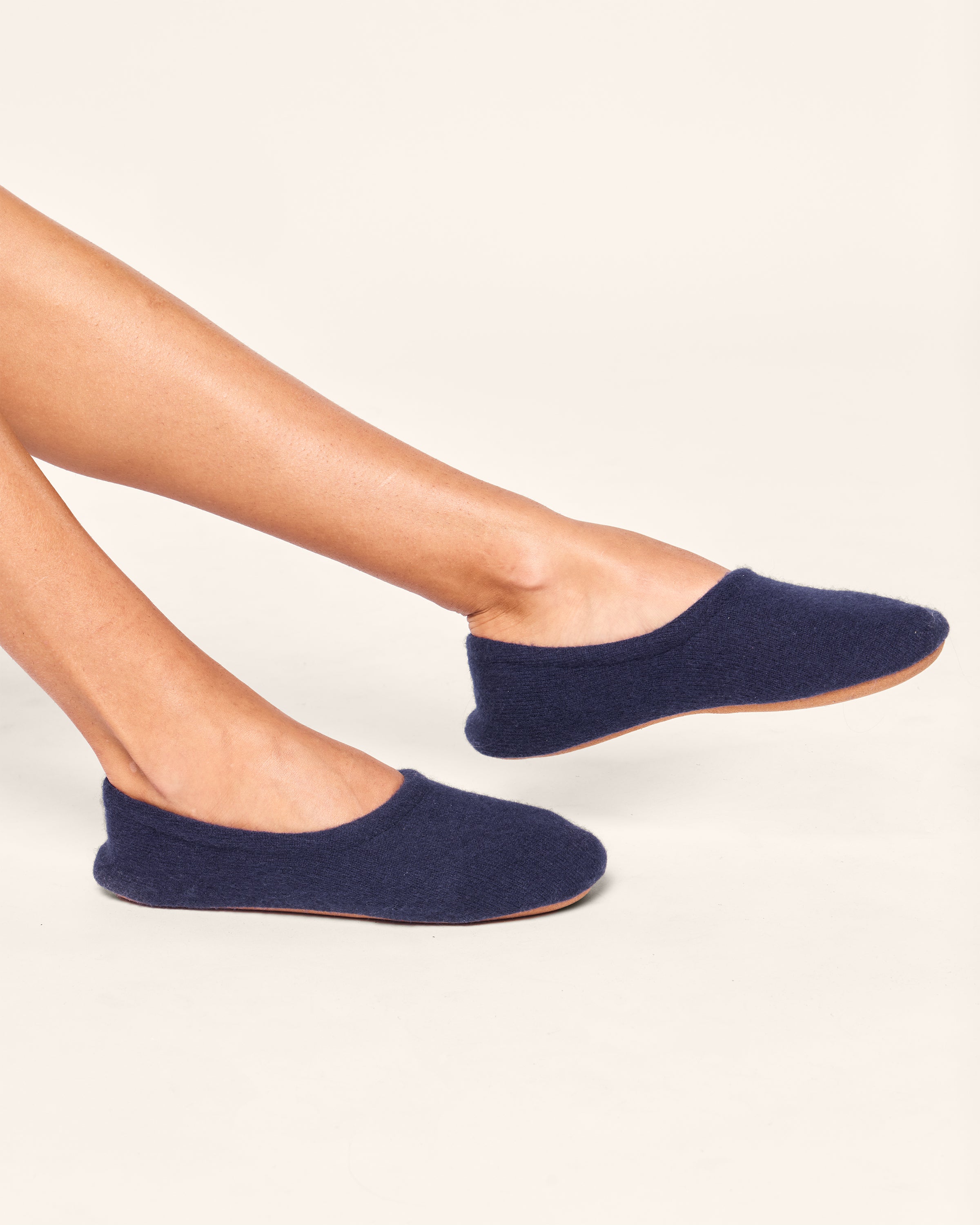 The lower legs wear Petite Plumes Womens Cashmere Slippers in Navy. These dark blue, soft slippers feature a simple design with a rounded toe and no visible fastenings, all set against a plain, light background. Crafted from Grade A pure cashmere for ultimate comfort.