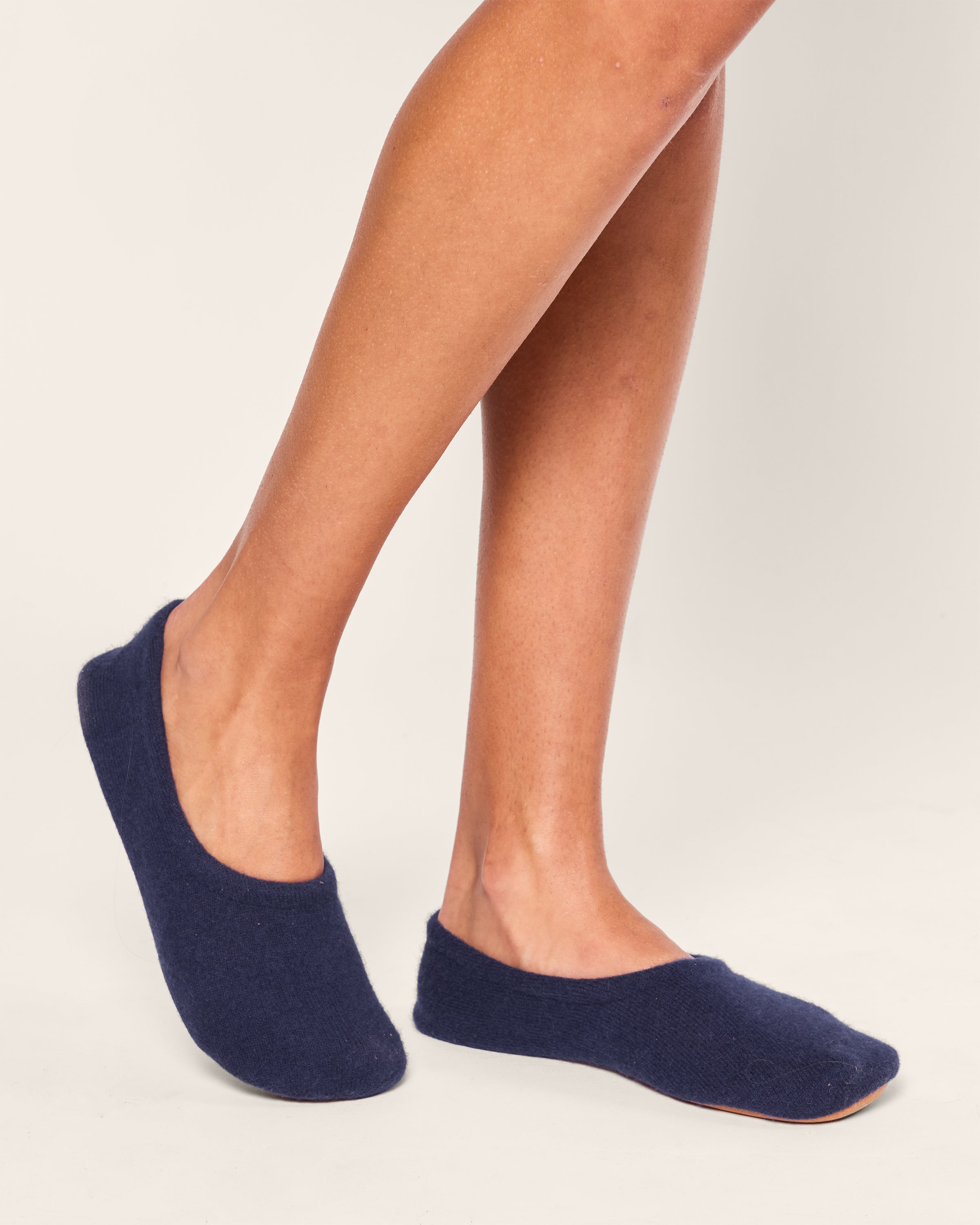 A person stands on a light surface wearing Womens Cashmere Slippers in Navy from Petite Plume, emphasizing the snug fit and simple design. The focus is on the legs and feet, reminiscent of a plush texture.