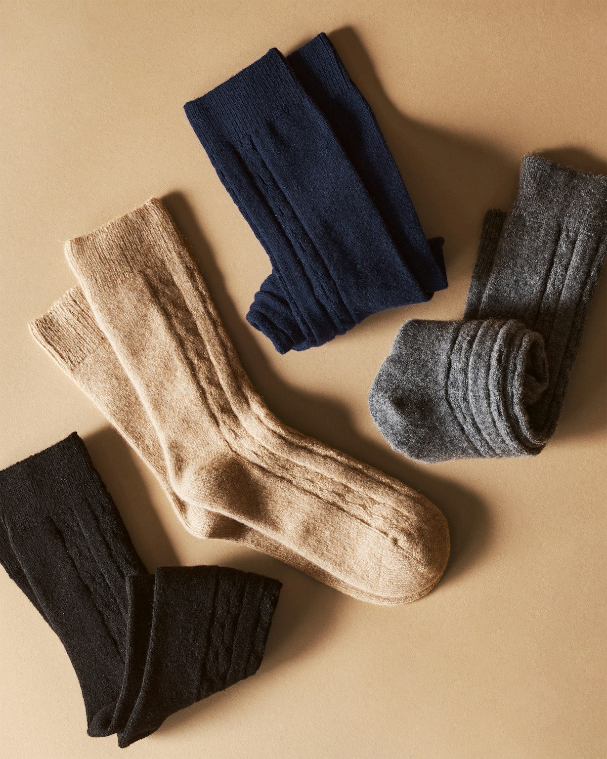 Four pairs of Petite Plume Mens Cashmere Socks rest on a beige surface, showcasing luxurious Grade A cashmere in navy blue, gray, beige, and black. Each is neatly folded to highlight their ribbed detail.