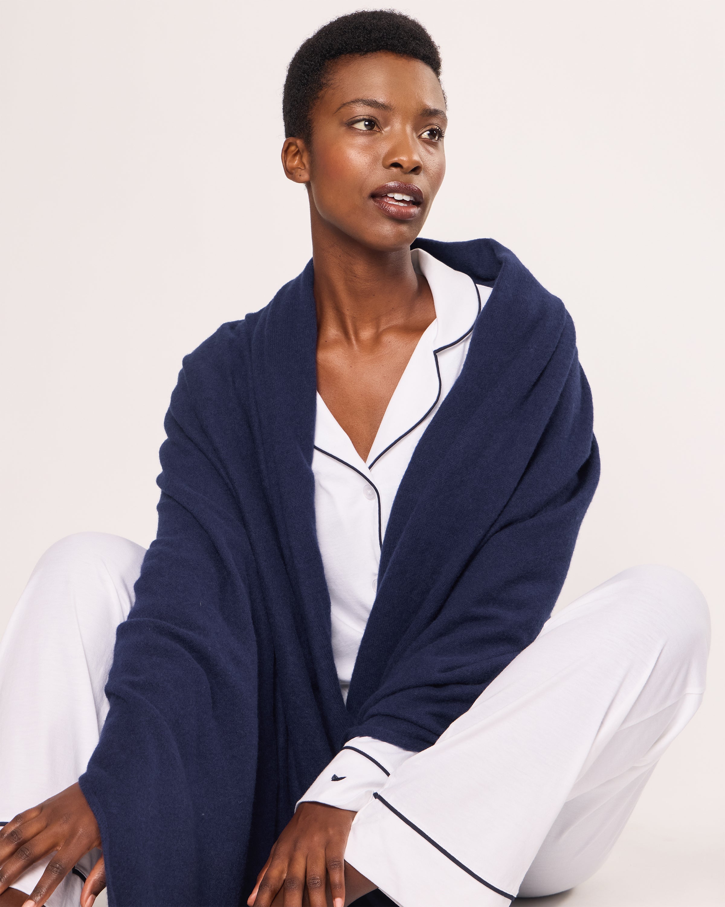 A person dons Petite Plumes Womens Cashmere Wrap in Navy and white pajamas with black piping, sitting calmly on a light surface. The plain white background highlights the elegance of the Grade A cashmere.