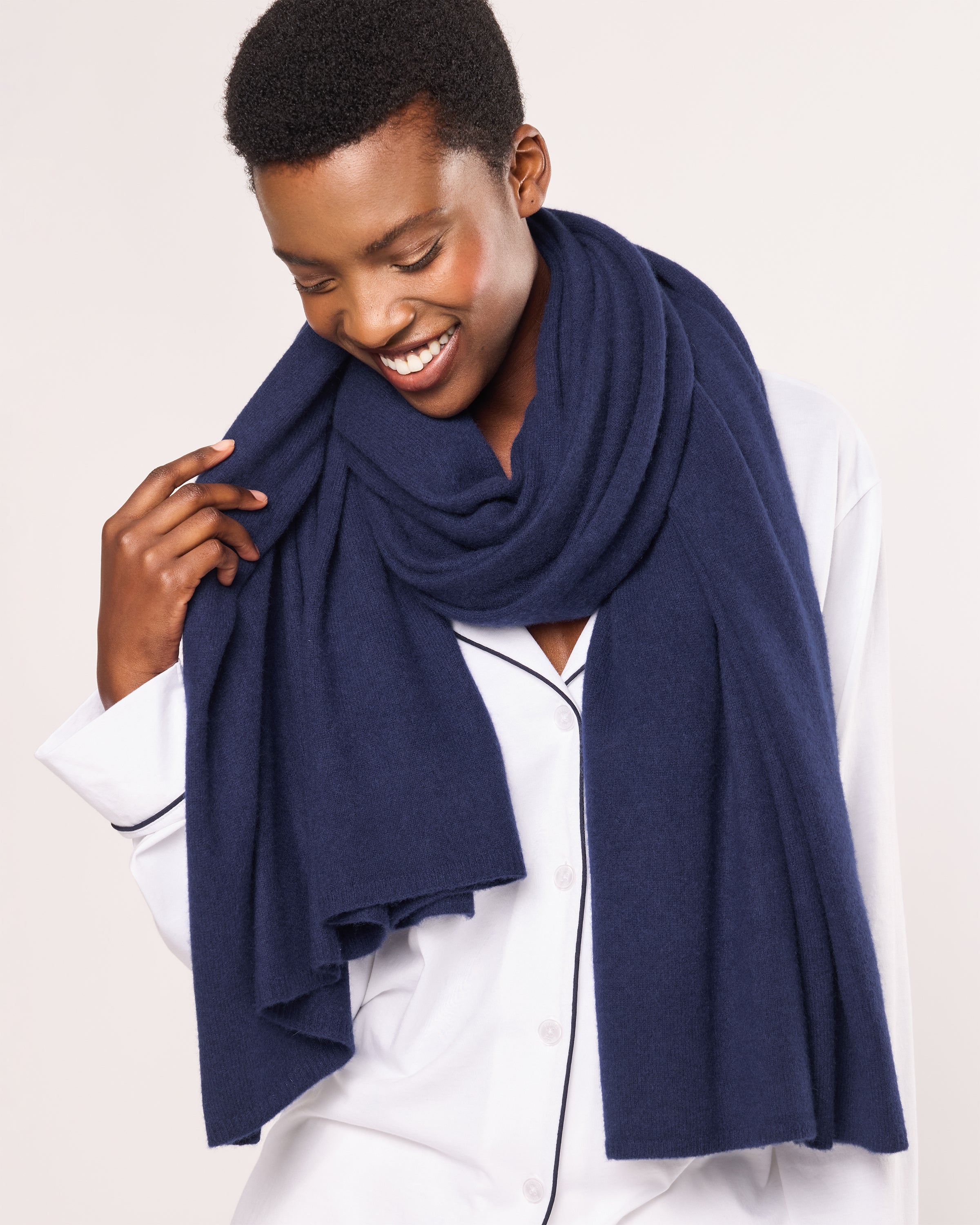 A joyful person smiles, draped in a Petite Plume Womens Cashmere Wrap in Navy. The luxurious Grade A cashmere wrap elegantly complements their light shirt, shining against the plain, light-colored backdrop.