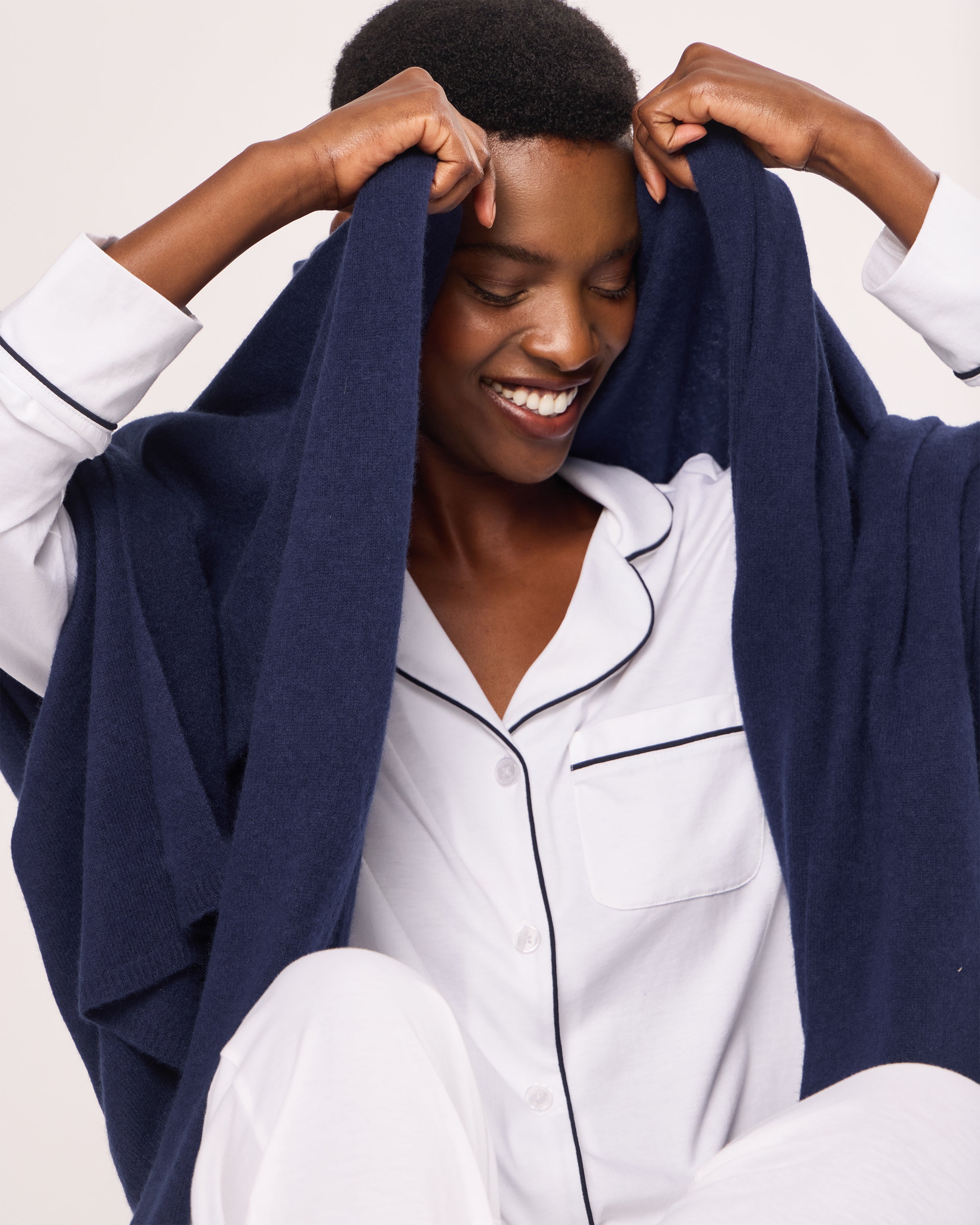 A person in a Petite Plume white pajama set with black piping smiles while draping themselves in a Womens Cashmere Wrap in Navy. The background is plain and light-colored.