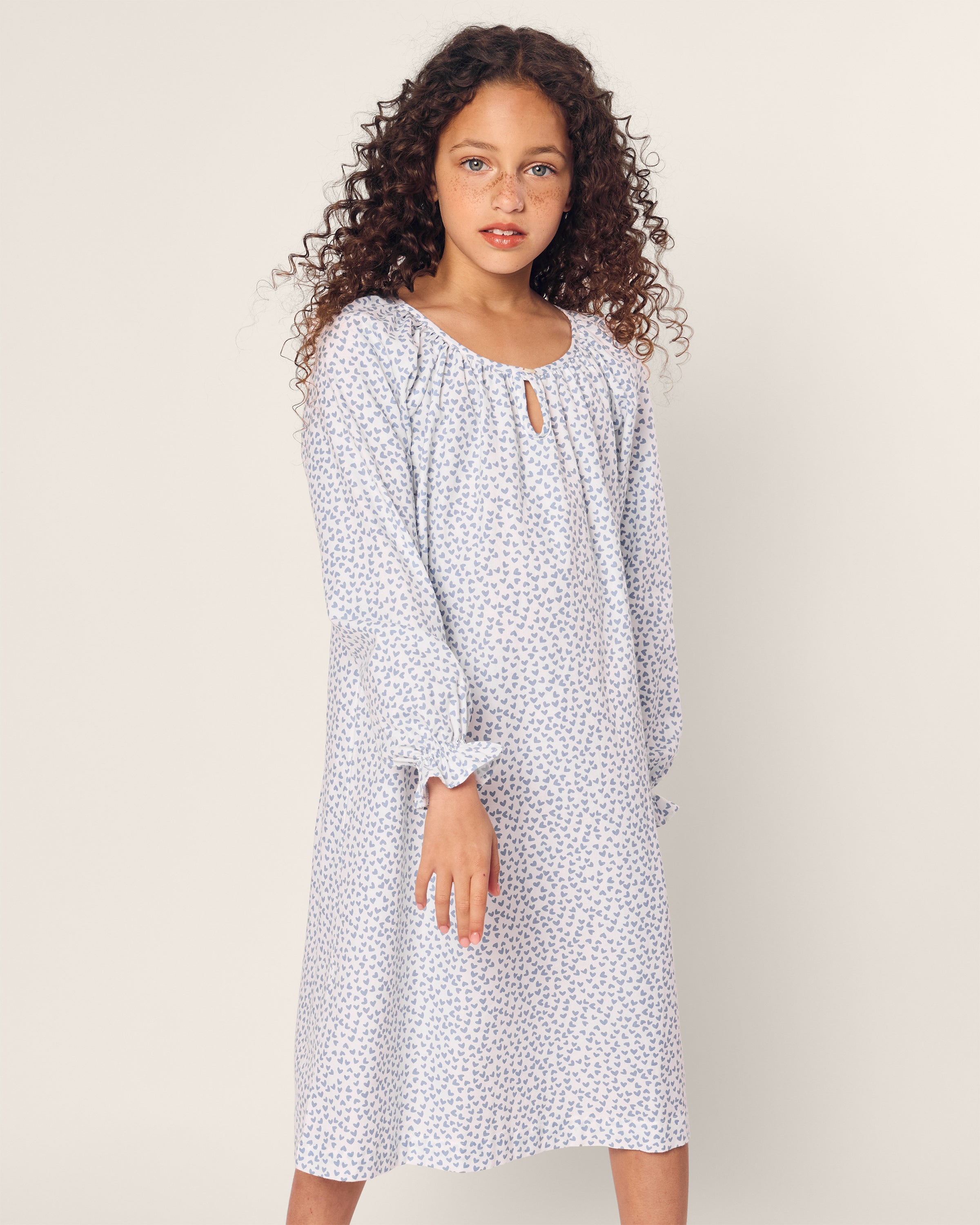 A young girl with curly hair confidently poses against a plain background, wearing Petite Plumes Girls Twill Delphine Nightgown in Bluehearts, featuring a light blue color and subtle floral pattern that adds elegance.