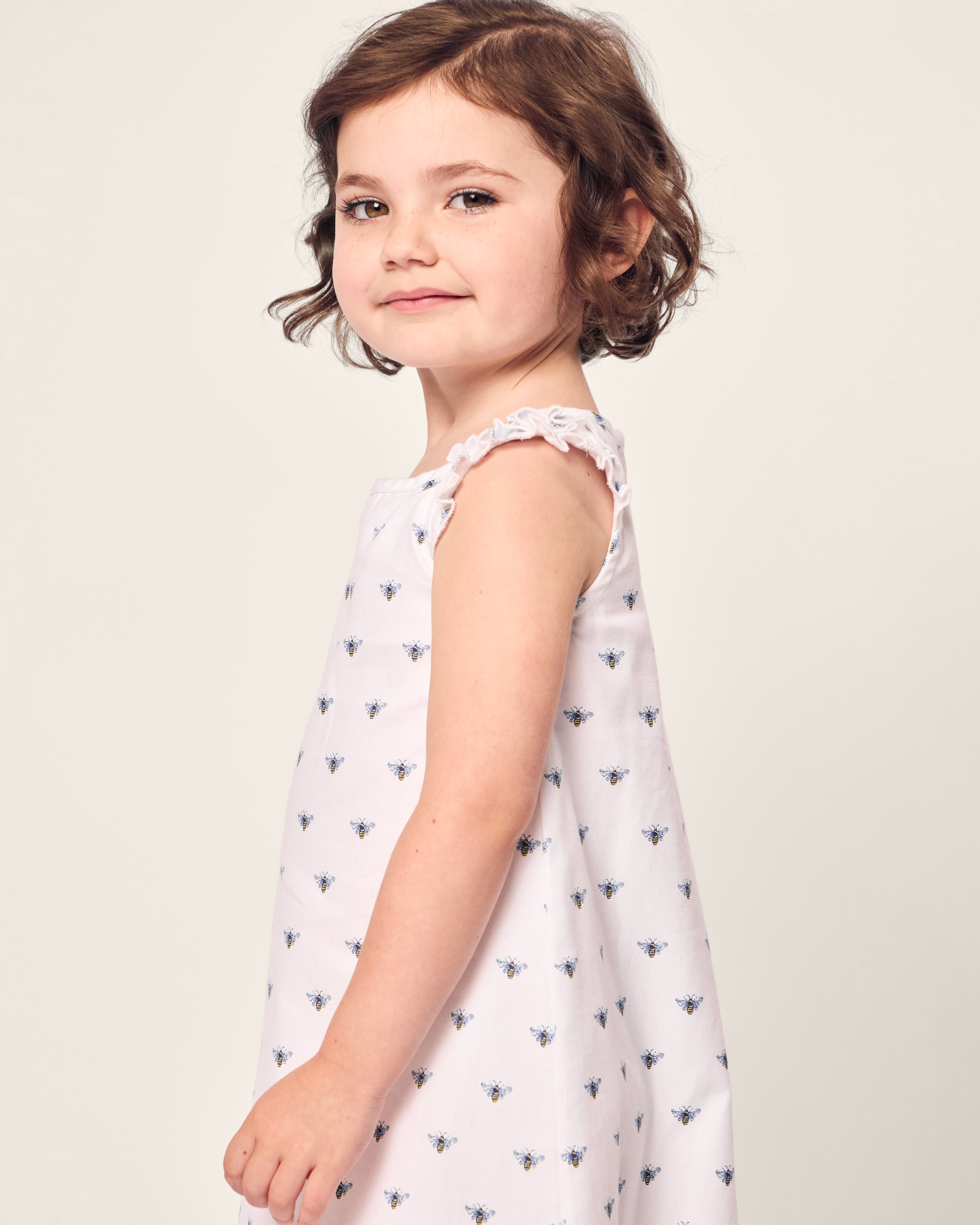 A young girl with a brown bob smiles slightly, wearing Petite Plumes Girls Twill Amelie Nightgown in Buzzing Bees. Her soft cotton gown features a white sleeveless design adorned with small bees, contrasting against the plain, light background.