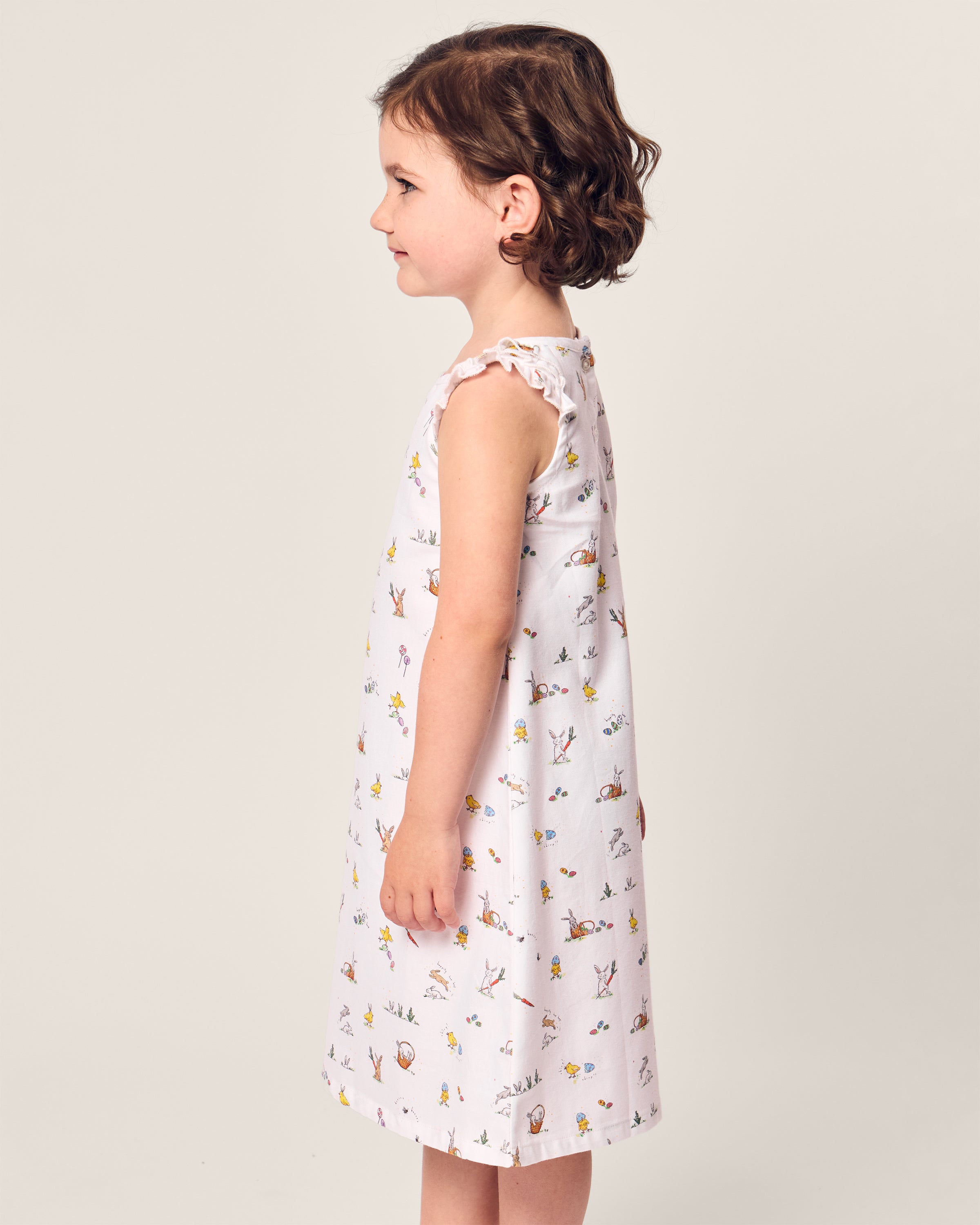 A child with short, curly brown hair models the Petite Plume Girls Twill Amelie Nightgown in Easter Frolic, showcasing its luxurious ruffled sleeves and vibrant patterns against a simple background.