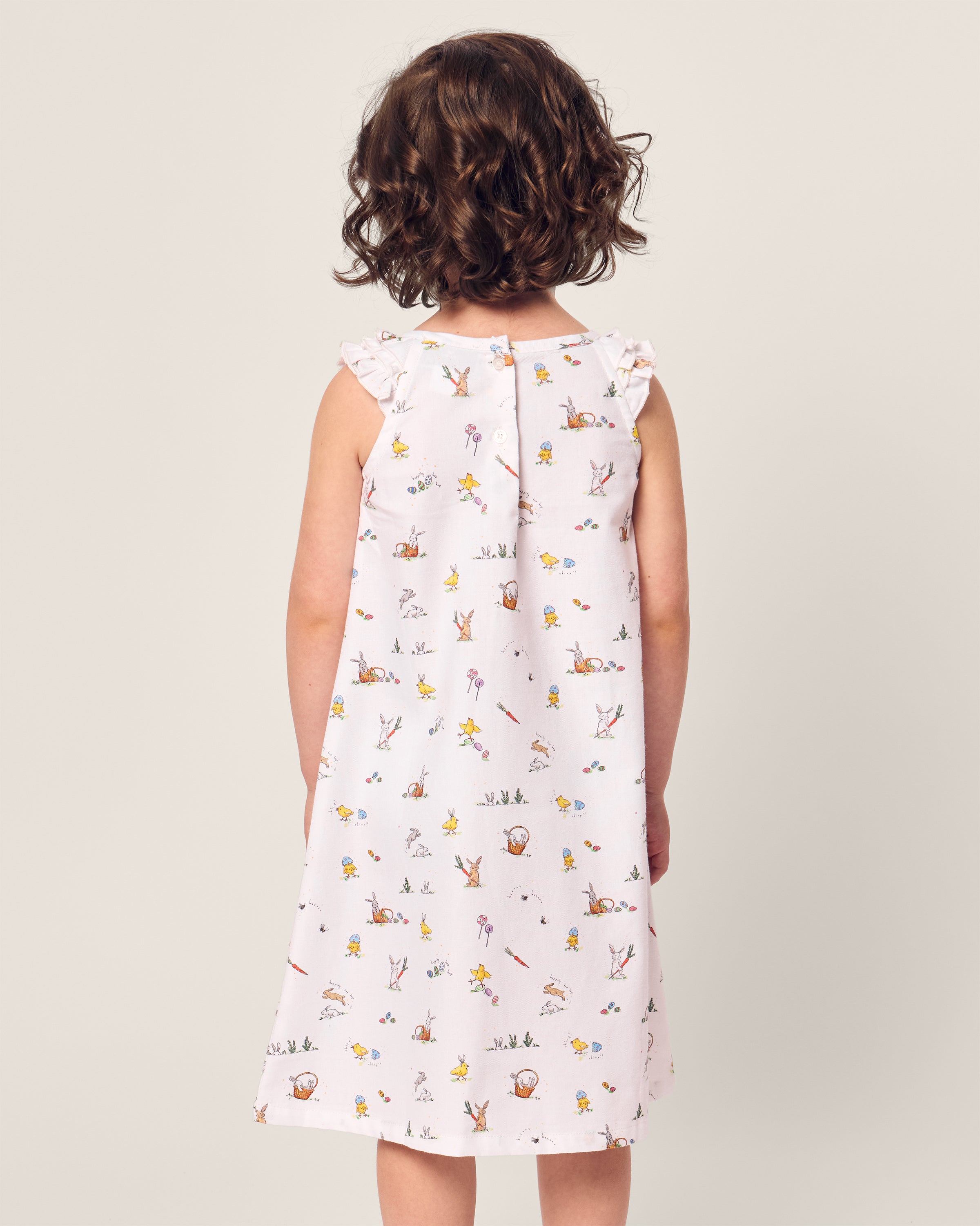 A young girl with curly hair stands facing away, wearing the sleeveless Girls Twill Amelie Nightgown in Easter Frolic by Petite Plume. The soft neutral background highlights her casual and whimsical attire.