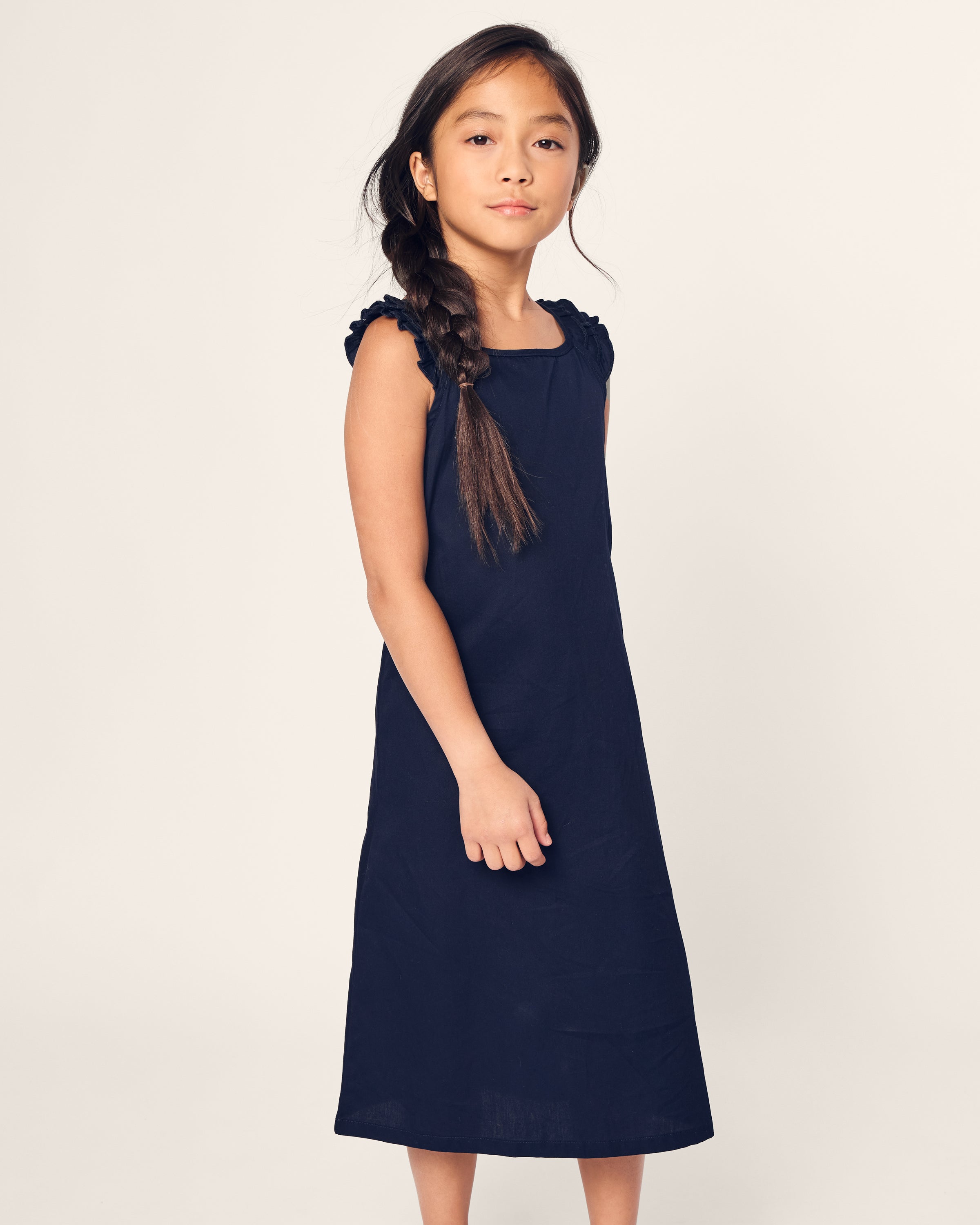 A young girl with long brown hair in a braid stands confidently against a plain light background, wearing Petite Plumes Girls Twill Amelie Nightgown in Navy. The sleeveless nightgown is made of high-quality cotton and features elegant ruffled shoulders.