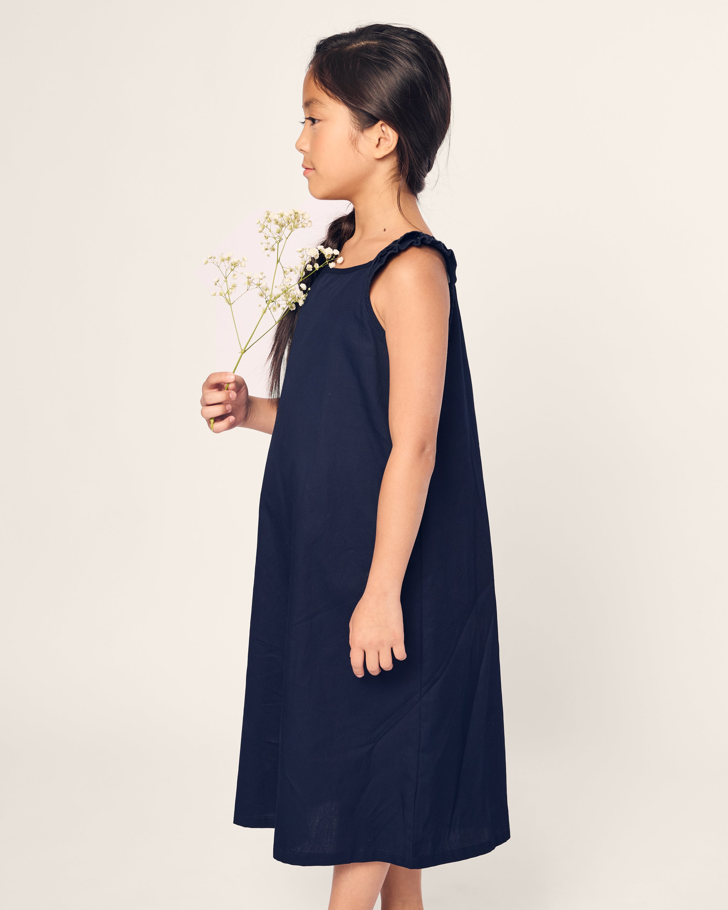 A young girl wearing Petite Plumes Girls Twill Amelie Nightgown in Navy, made from quality cotton, stands in profile against a light background holding small white flowers. Her dark hair is tied back as she looks forward with a calm expression.