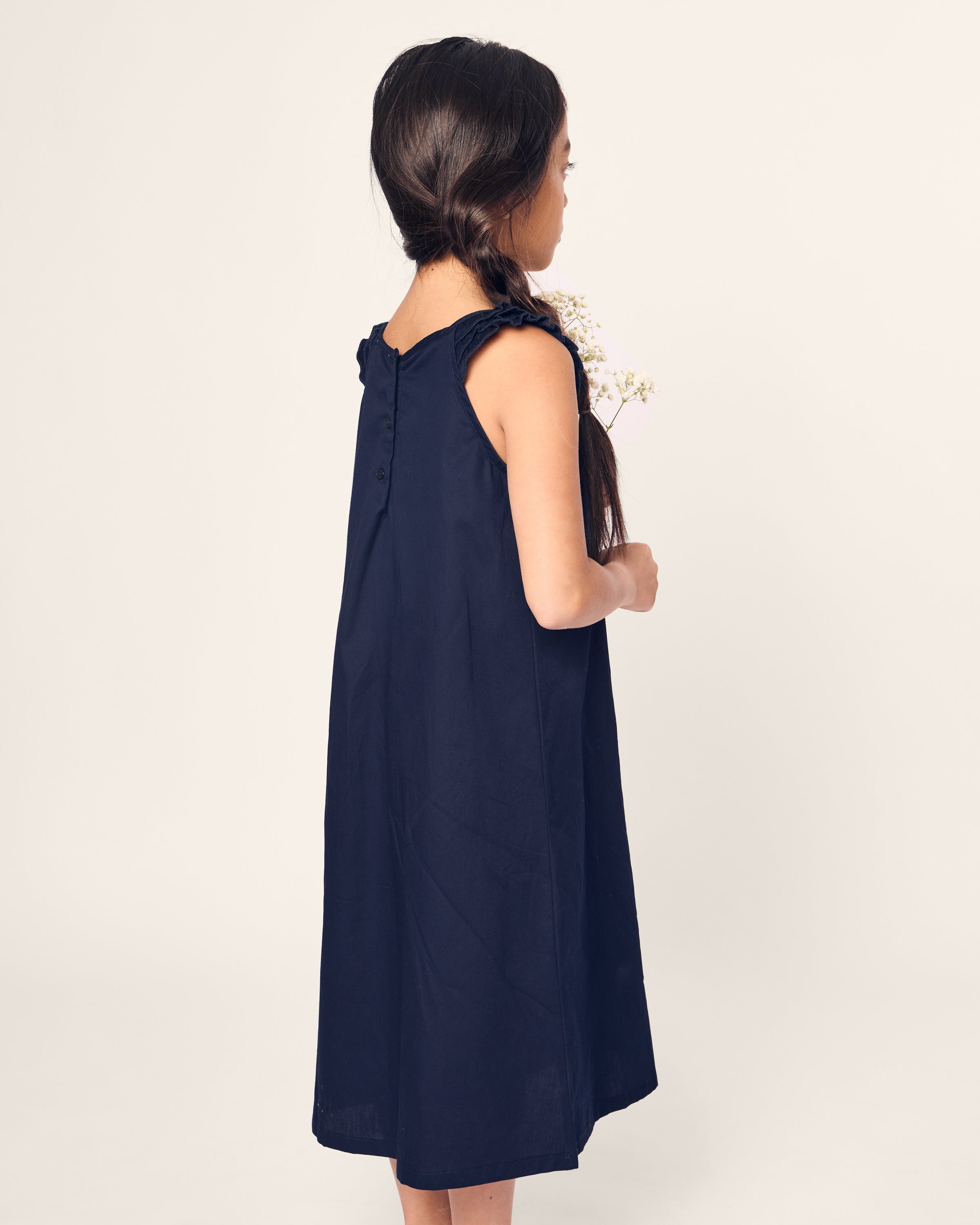 A young girl with long hair stands facing left, holding small white flowers while wearing the Petite Plume Girls Twill Amelie Nightgown in Navy. The sleeveless dress is made of fine cotton with ruffled shoulders, set against a plain light-colored background highlighting her elegant silhouette.