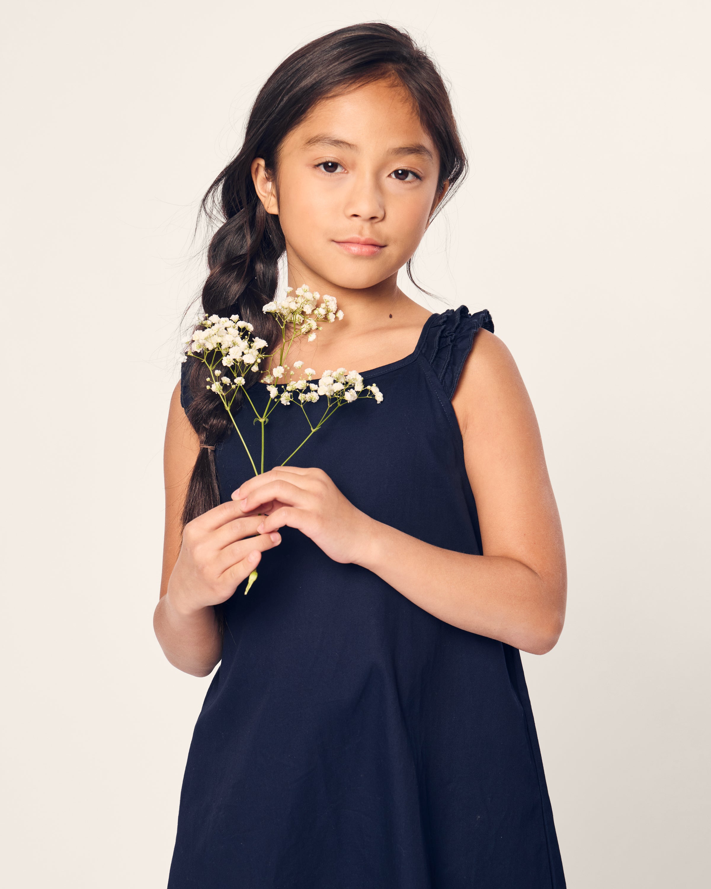 A young girl with long, dark hair in a braid wears the Petite Plume Girls Twill Amelie Nightgown in Navy and holds small white flowers against a plain light background, looking at the camera.