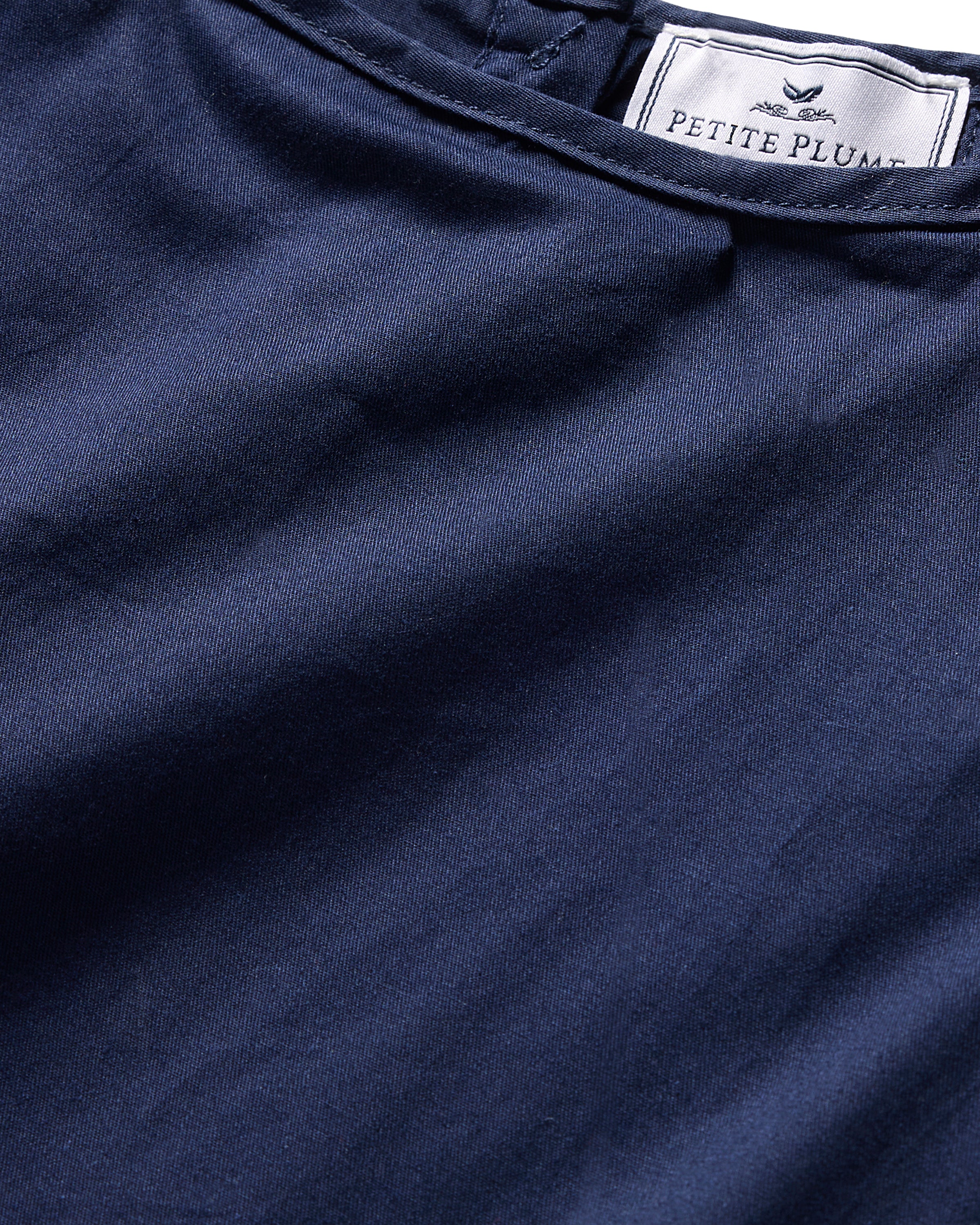 A close-up of the navy twill fabric from the Girls Twill Amelie Nightgown shows a PETITE PLUME label with a feather design, suggesting luxurious quality. The textured cotton is soft to touch, ideal for cozy sleepwear.