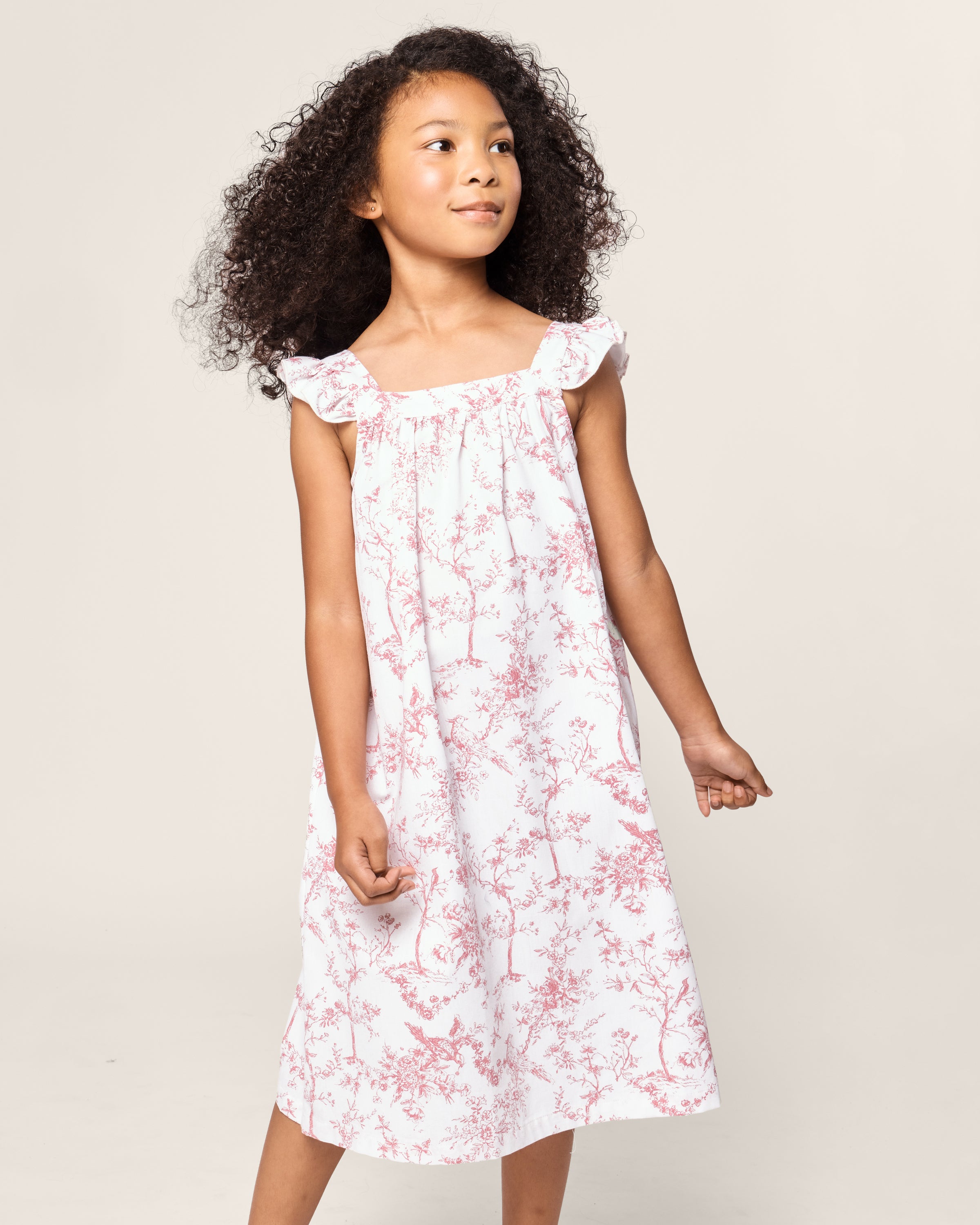 A young girl with curly hair wears the Petite Plume Girls Twill Clara Nightgown in Pink Timeless Toile. She stands against a plain background, looking slightly to the side, with a calm expression.