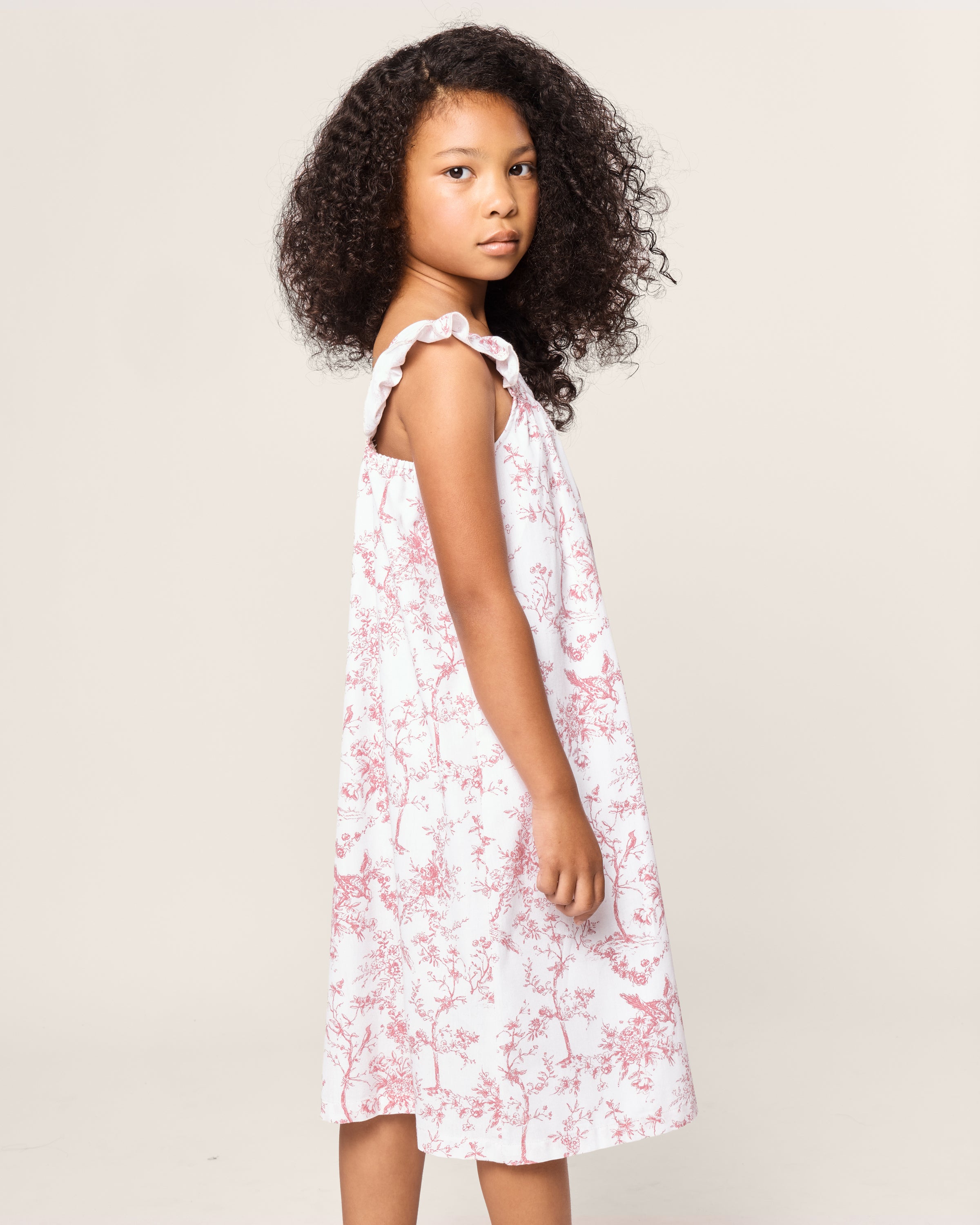 A young girl with curly hair, wearing Petite Plumes Girls Twill Clara Nightgown in Pink Timeless Toile, stands slightly sideways. She gazes directly at the camera against a plain background.