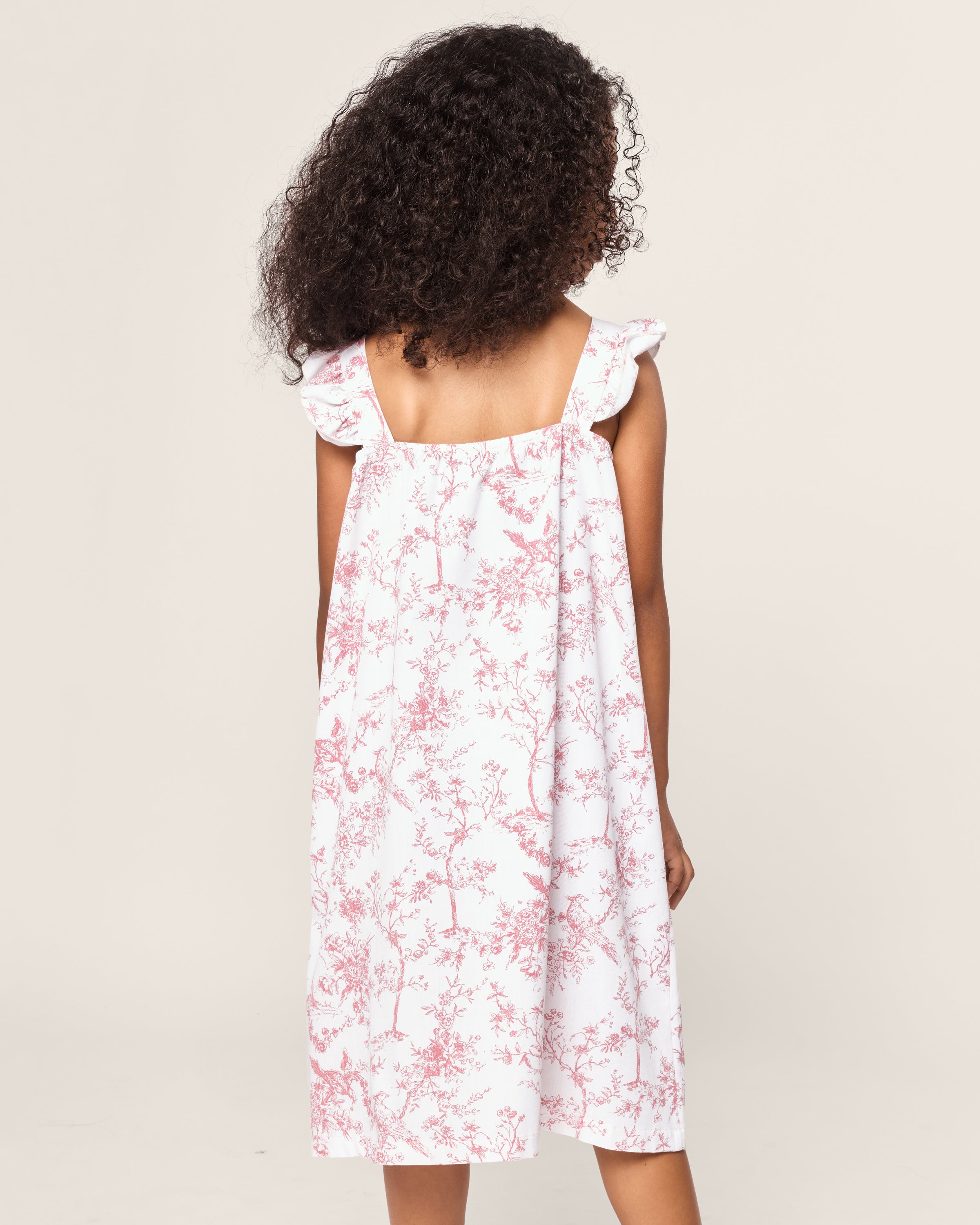 A girl with curly hair faces away, wearing Petite Plumes Girls Twill Clara Nightgown in Pink Timeless Toile, featuring delicate botanical flowers and ruffled shoulder straps. The background is plain off-white.