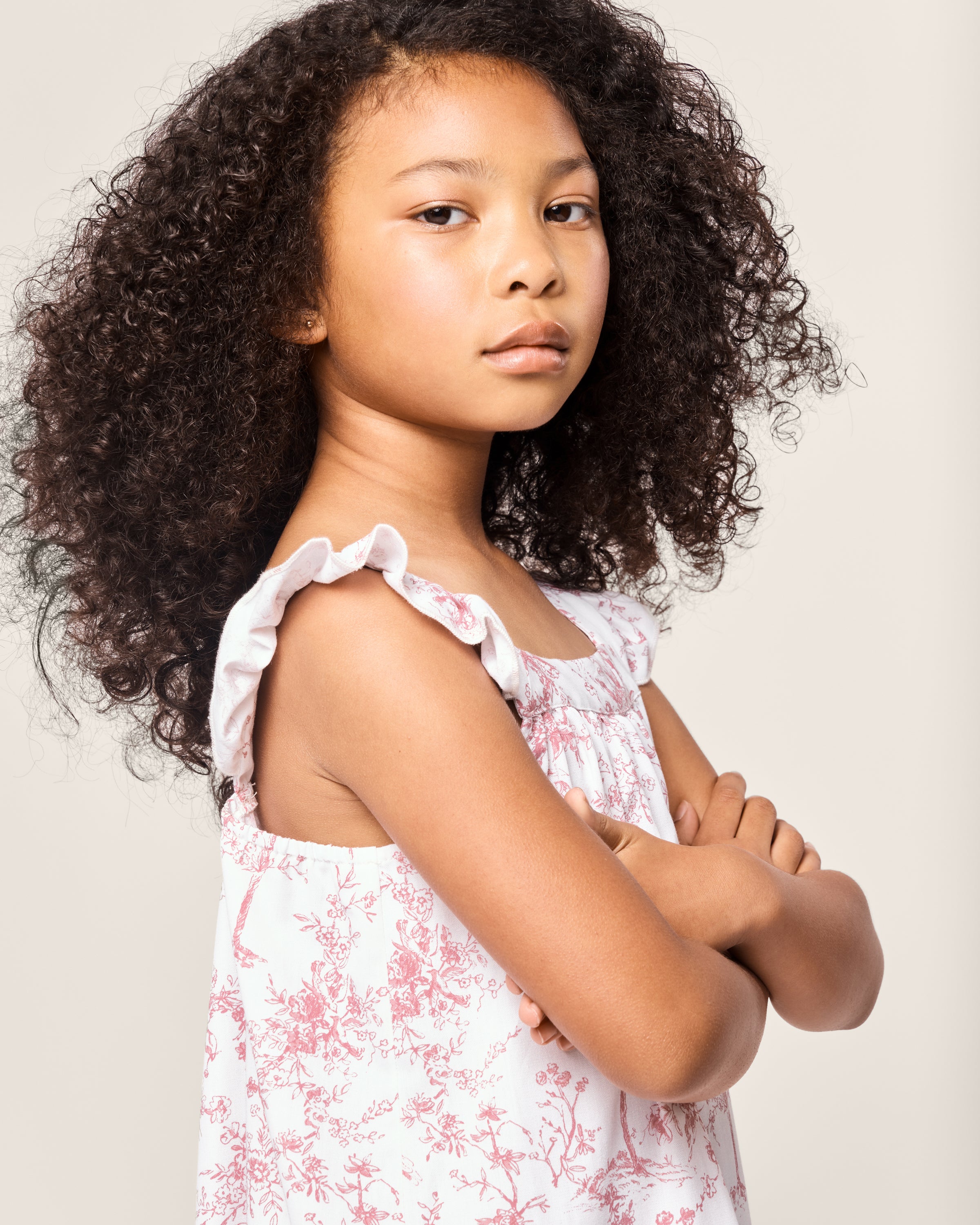 A young girl with curly hair stands confidently, arms crossed, in the Girls Twill Clara Nightgown in Pink Timeless Toile by Petite Plume. She gazes forward against a plain background, exuding youthful charm akin to luxury sleepwear.