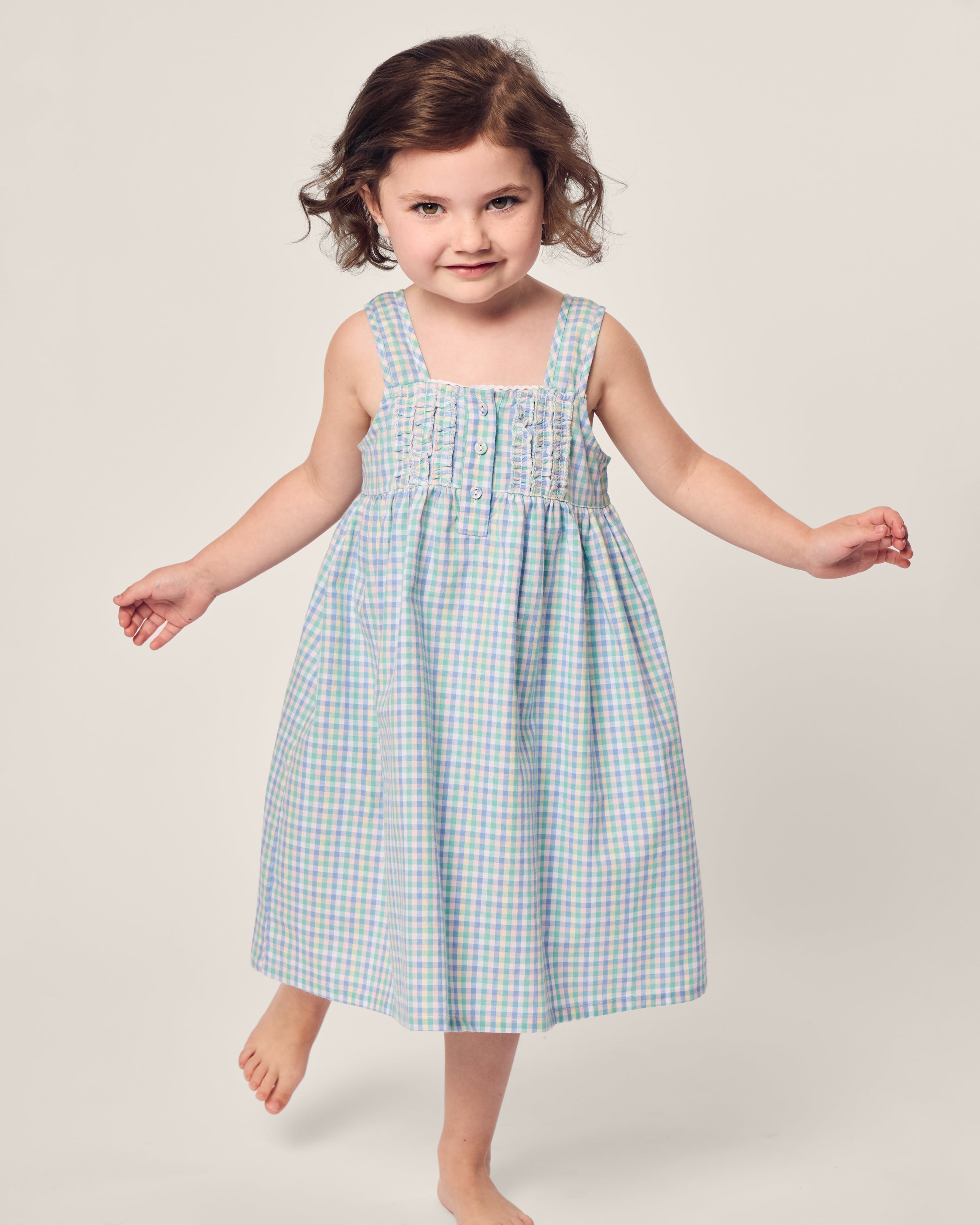 A curly-haired girl stands barefoot against a plain background, wearing Petite Plumes Girls Twill Charlotte Nightgown in Spring Gingham, with wide straps and a smocked bodice. Her arms are slightly outstretched as she gazes forward.