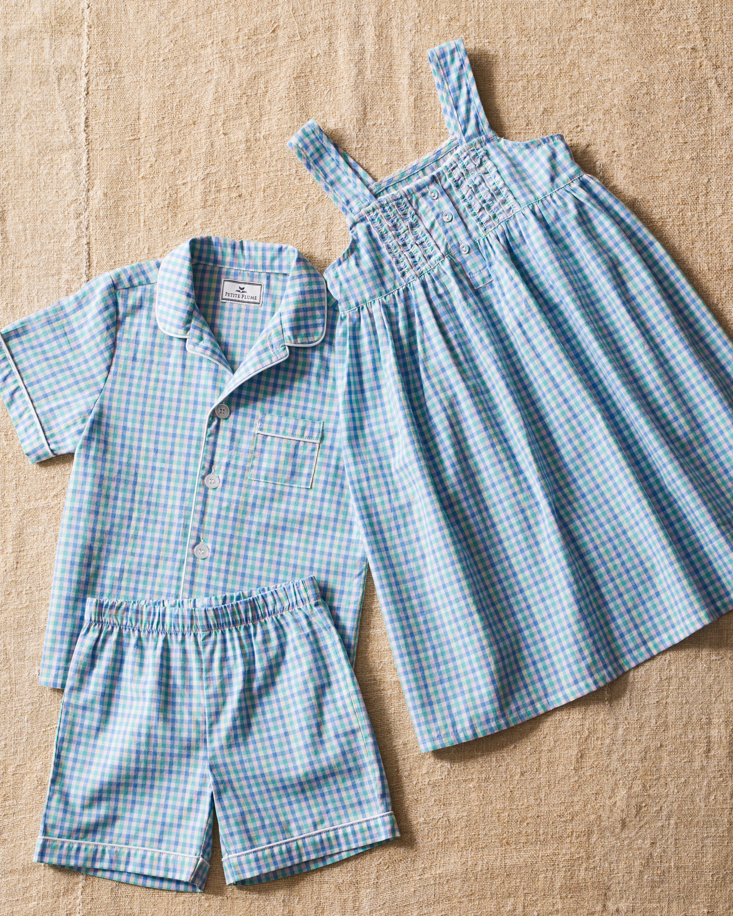 A set of childrens clothing on a beige surface includes a blue and green gingham print short-sleeve button-up shirt with matching shorts, and Petite Plumes Girls Twill Charlotte Nightgown in Spring Gingham featuring a sleeveless design and button details on the top.