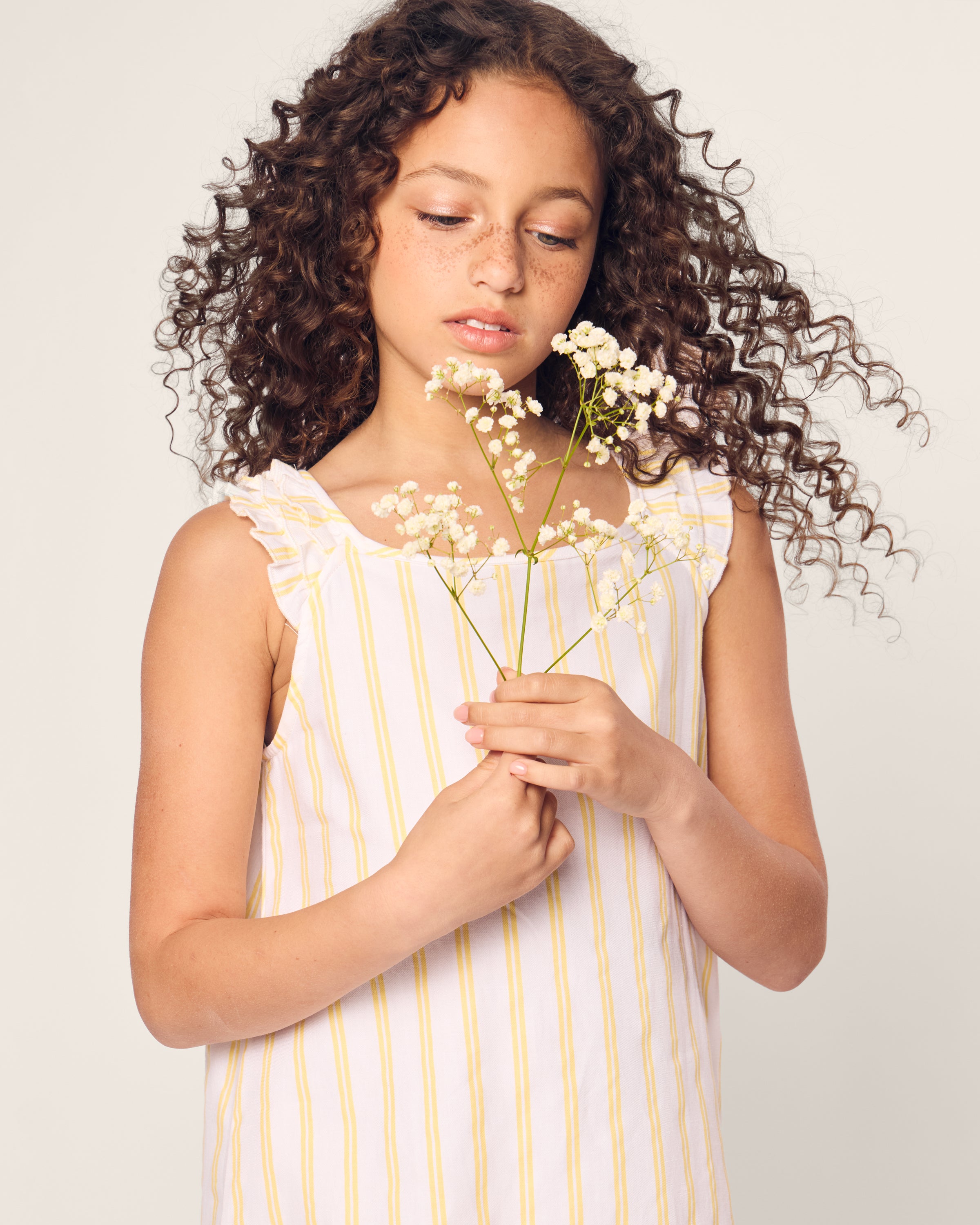 A freckled girl with curly brown hair holds small white flowers, wearing Petite Plumes Girls Twill Amelie Nightgown in Sunny Stripe. The background is a plain light gray.