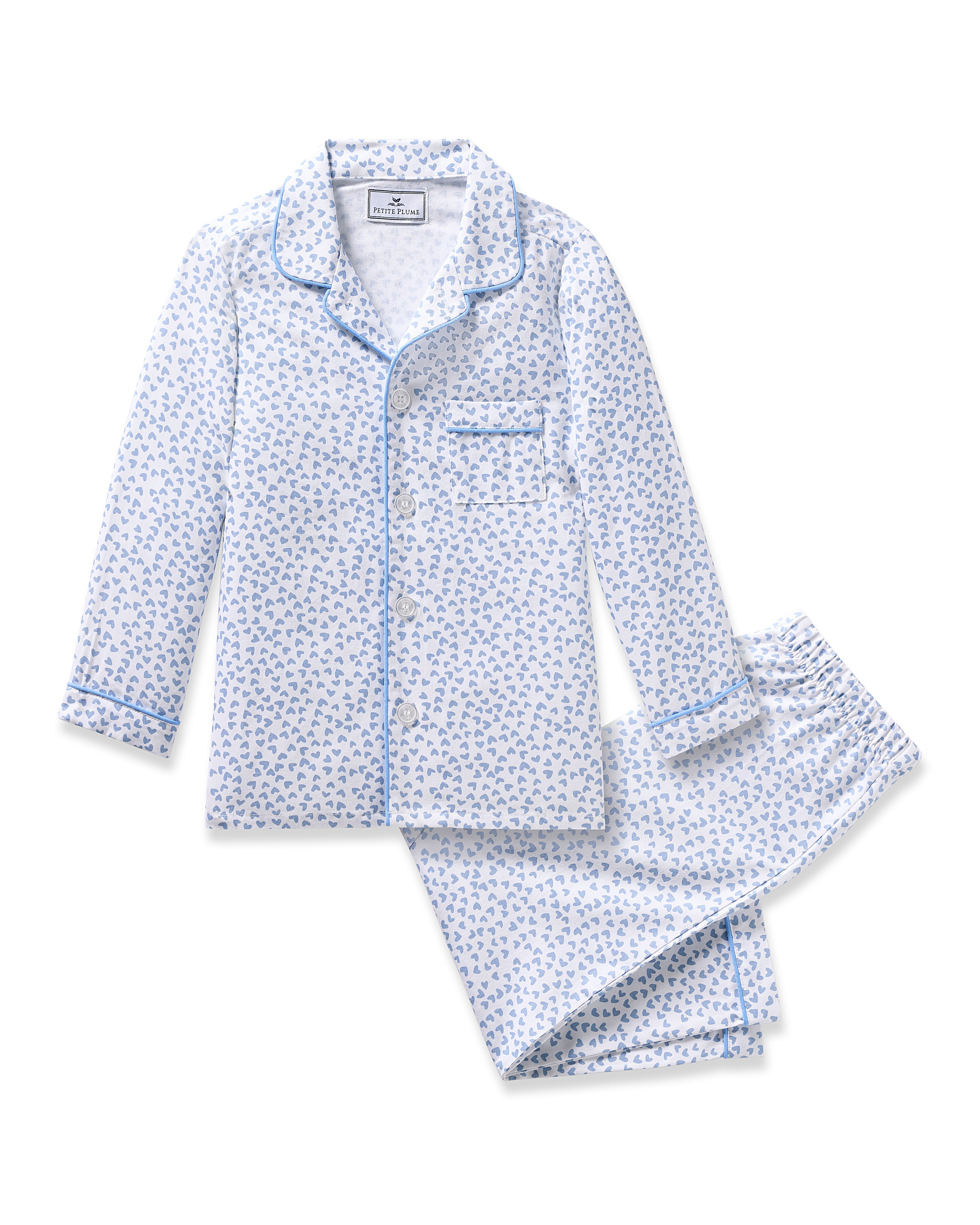 The Kids Twill Pajama in Bluehearts by Petite Plume is crafted from yarn-dyed cotton and adorned with a white fabric featuring a blue heart pattern. The top has a collar, long sleeves, and one chest pocket. The pants feature an elastic waistband. Both pieces are trimmed with blue piping for charm.