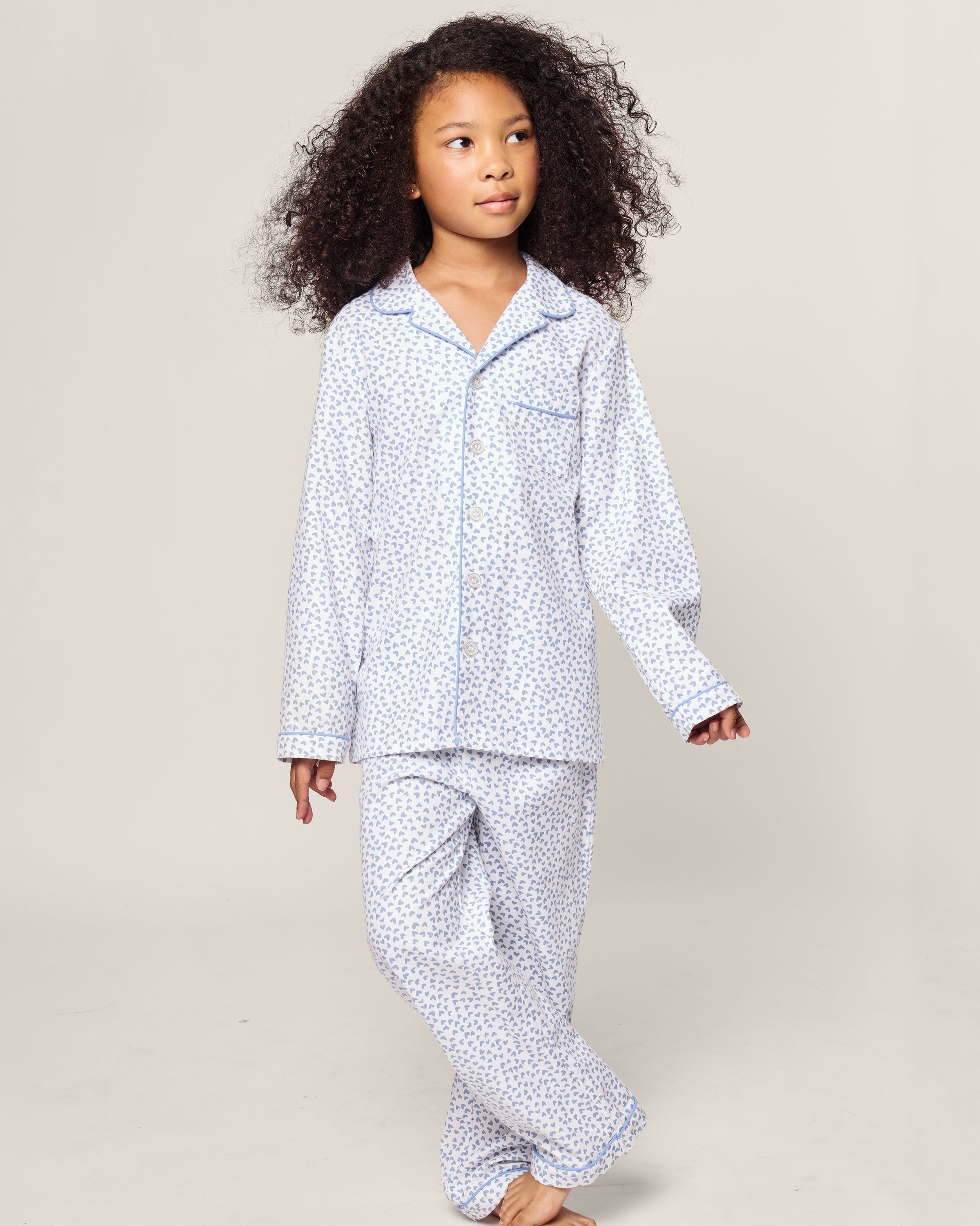 A child with curly hair wears Petite Plumes Kids Twill Pajama in Bluehearts, a light-hued yarn-dyed cotton set featuring small blue patterns. Standing barefoot against a plain background, the child looks slightly to the side.